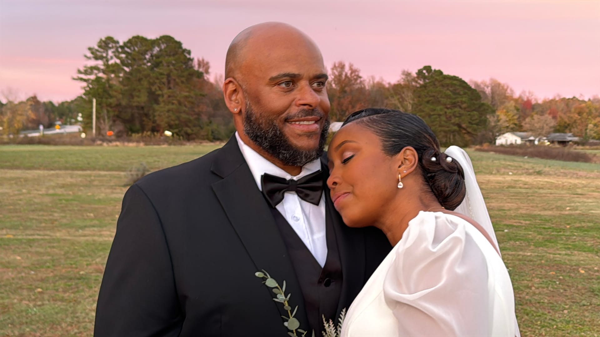 Bride in white gown rests her head on grooms shoulder as he smiles warmly while the two stand in an open farmland field beneath a soft pink sunset sky at Scarborough House.