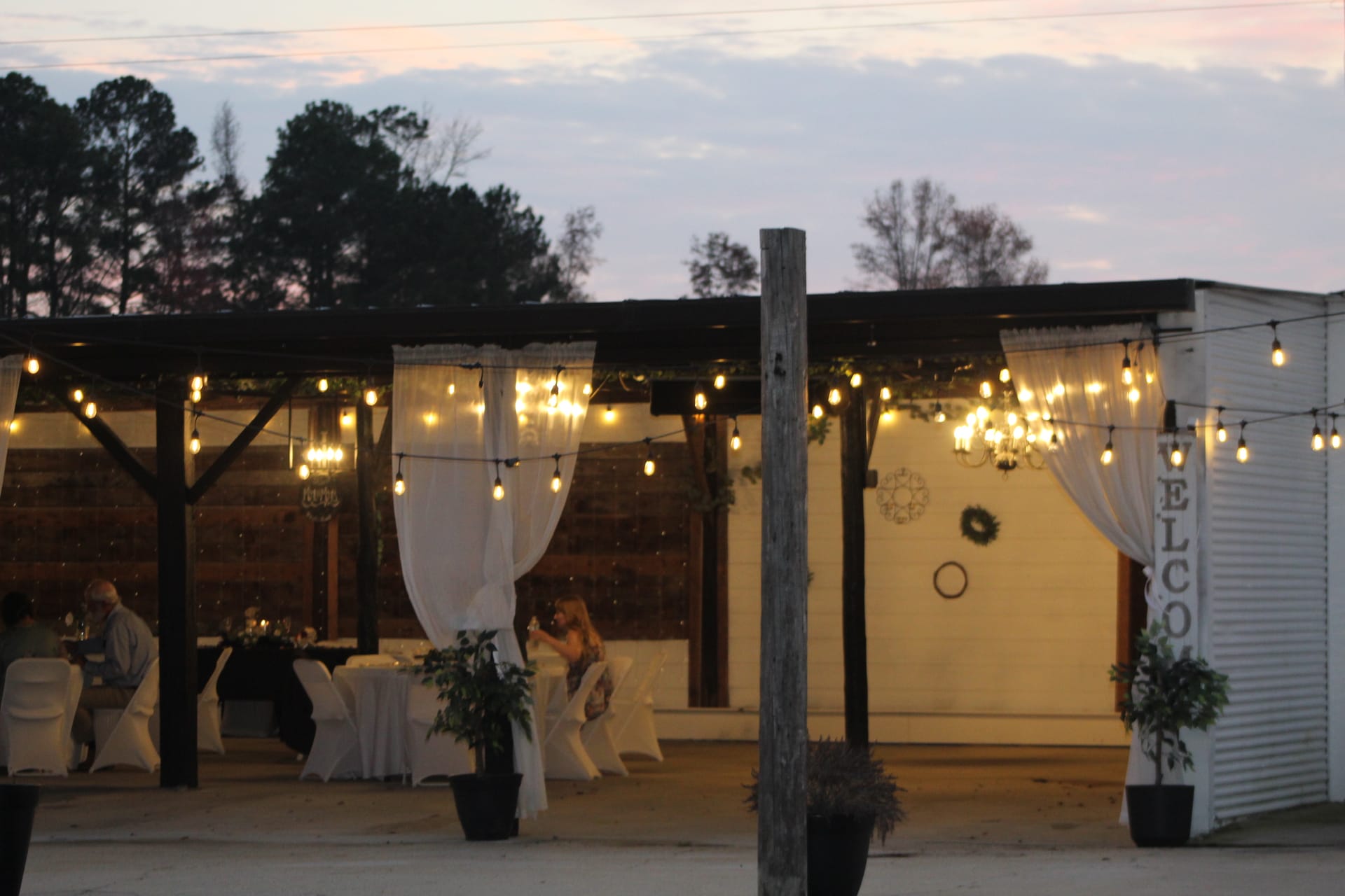 Guests seated at elegantly draped tables under a warmly lit open-air pavilion strung with Edison bulbs at dusk at Scarborough House wedding venue.
