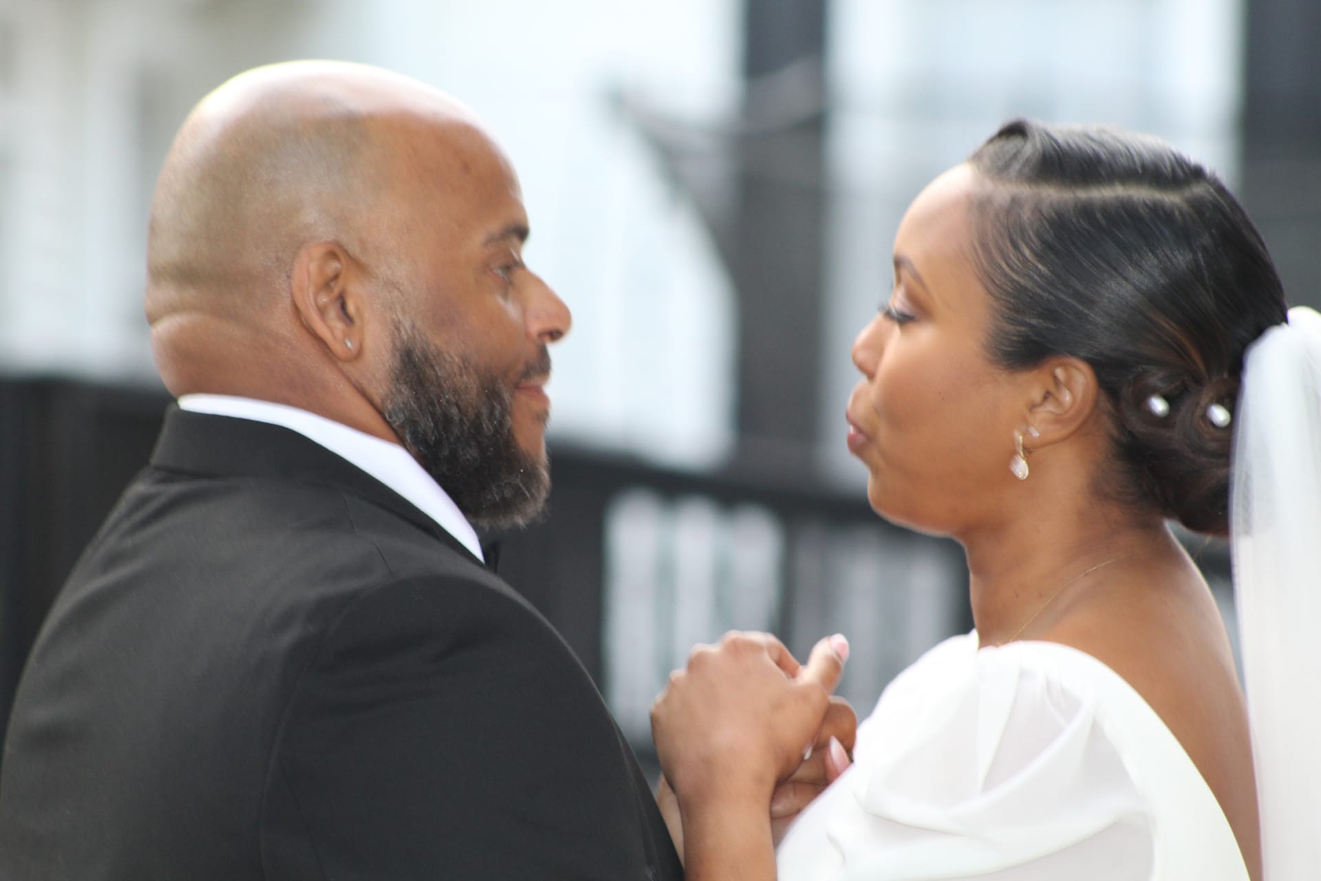 Bride in a white off-shoulder gown puckers for a kiss while holding hands with her tuxedo-clad groom during an intimate first-look moment at their wedding.