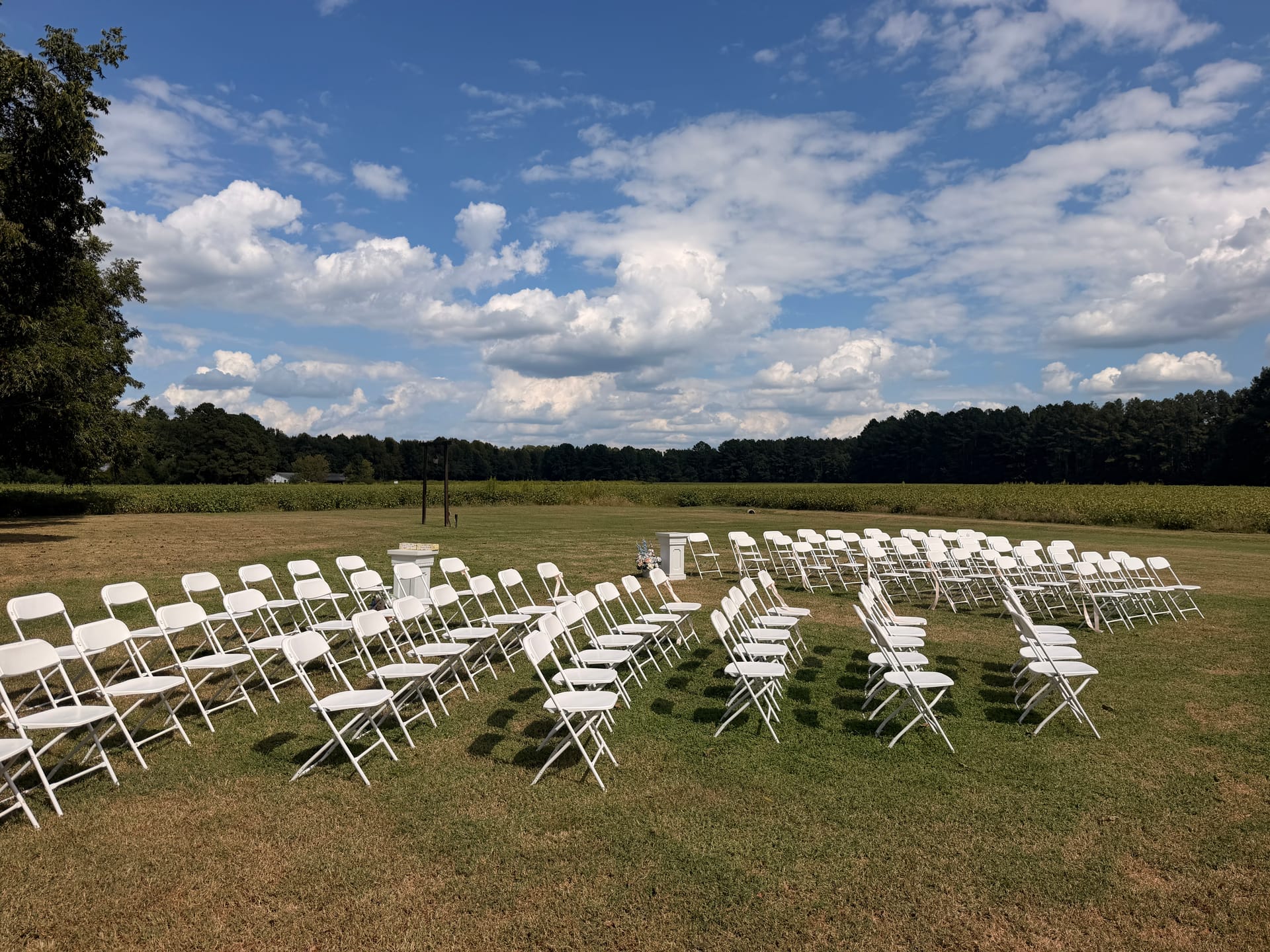 Rows of white folding chairs arranged on a sunlit lawn for an outdoor wedding ceremony at Scarborough House with a dramatic cloudy sky and farmland fields in the background.