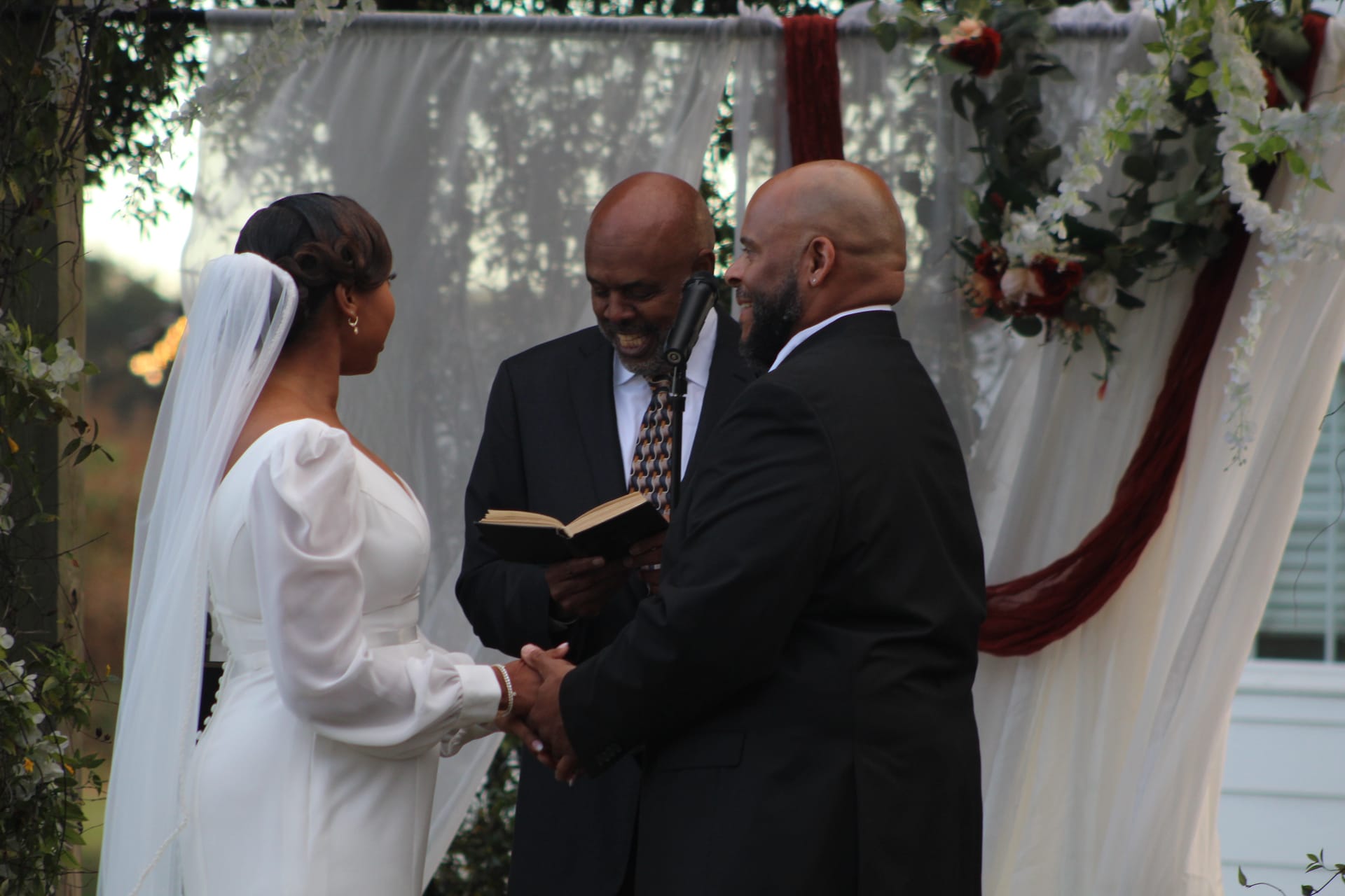 Bride in a white long-sleeve gown and groom in a black suit hold hands during their outdoor wedding ceremony beneath a floral-draped arch with burgundy and white decor at Scarborough House.