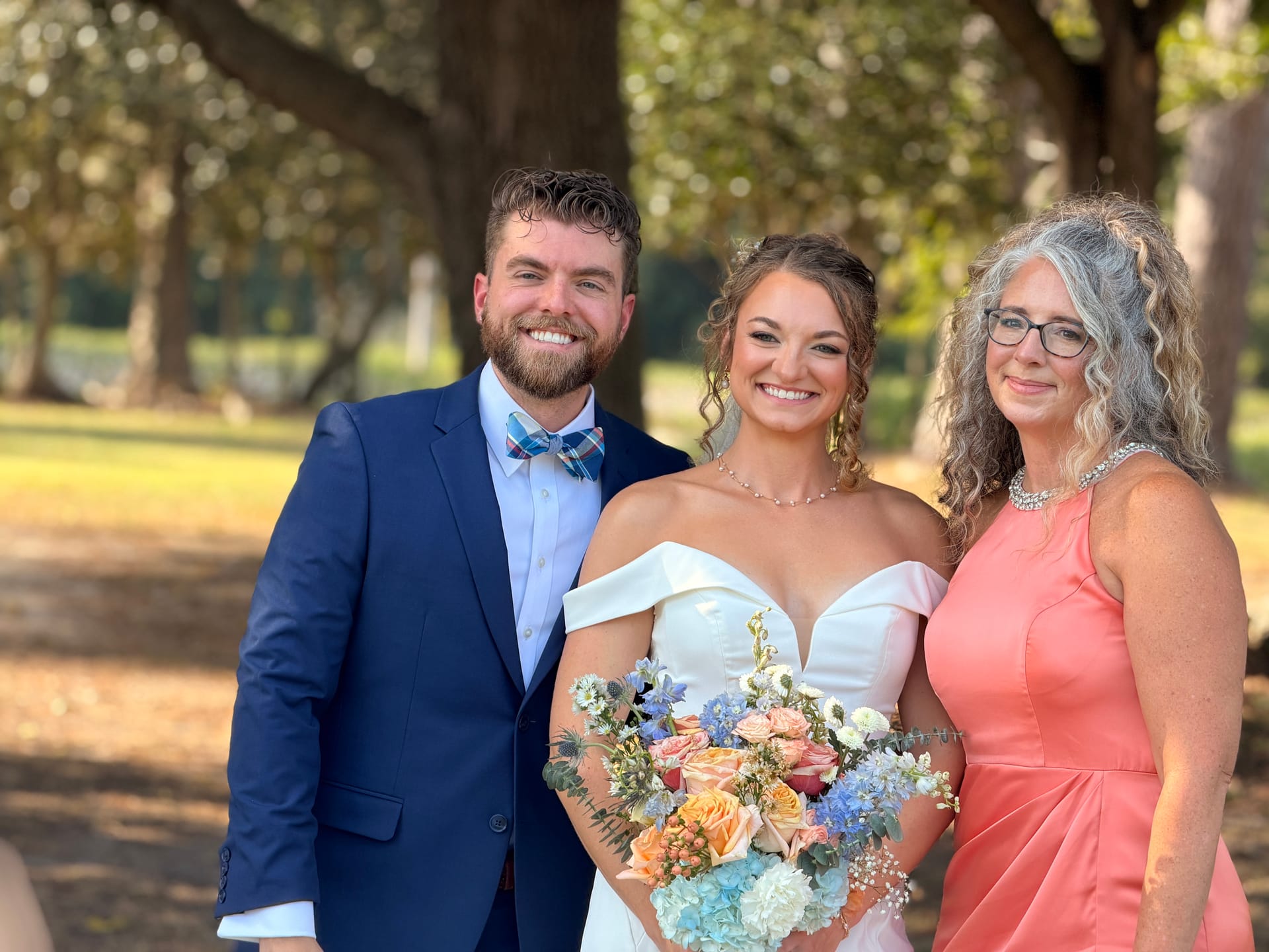 Bride in an off-shoulder white gown holding a pastel bouquet stands between the groom in a navy suit with plaid bow tie and an older female guest in coral, all smiling beneath the sweeping oak trees at Scarborough House.