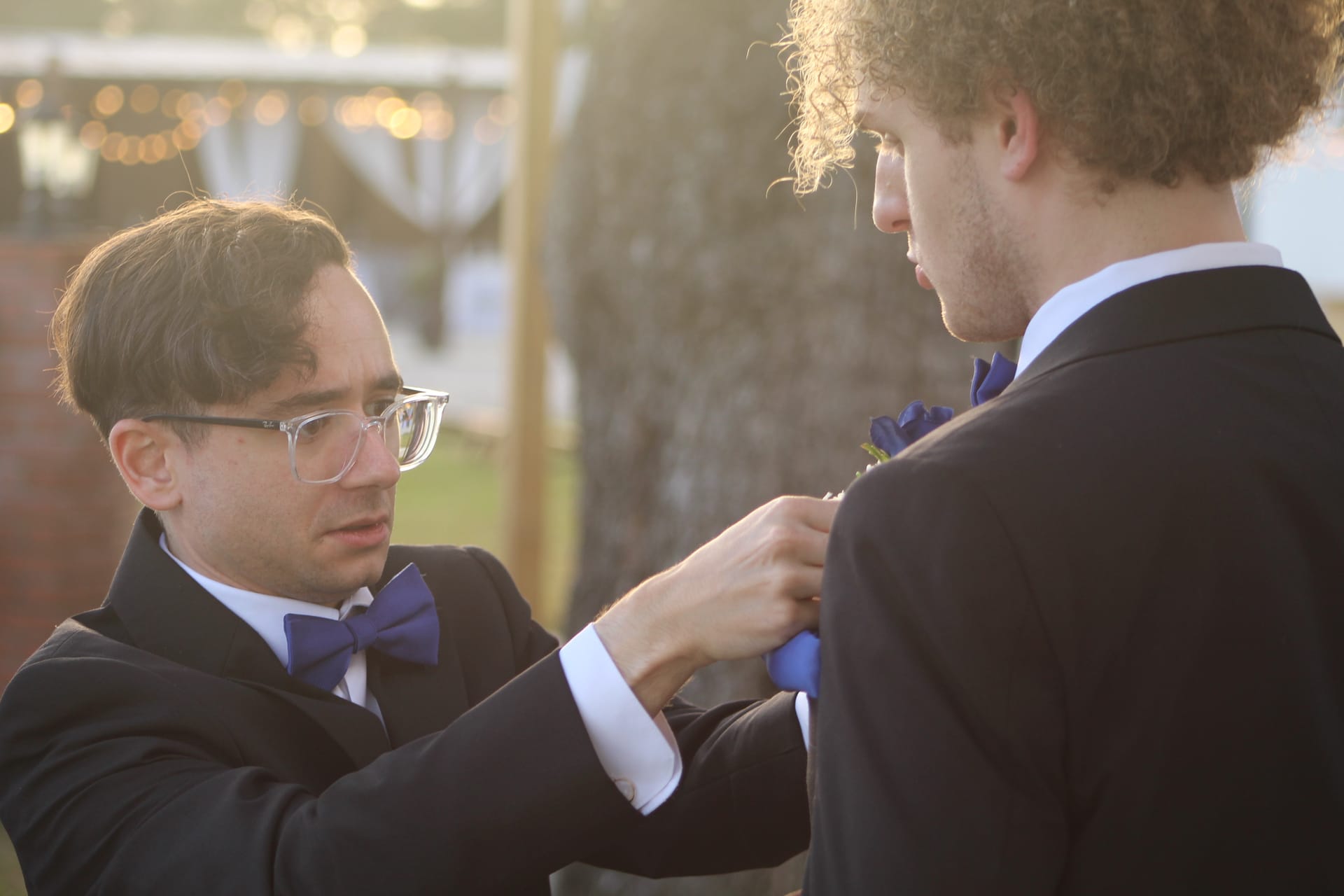 A groomsman in a black tuxedo and purple bow tie carefully pins a blue boutonniere on another groomsman outdoors in warm golden-hour light at Scarborough House.