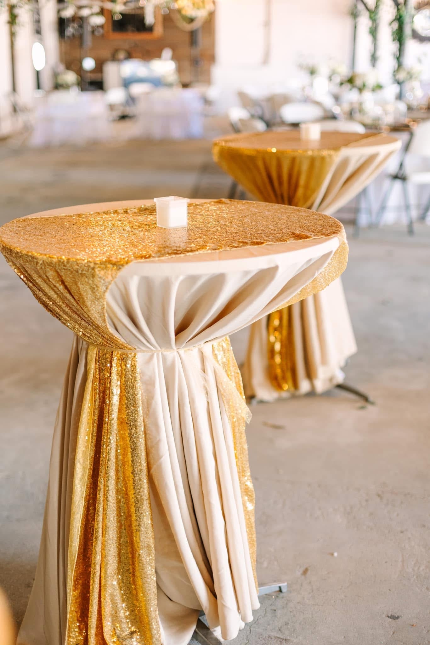 Row of tall cocktail tables draped in shimmering gold sequin and ivory satin linens topped with white votive candles inside the grand hall at Scarborough House wedding venue.