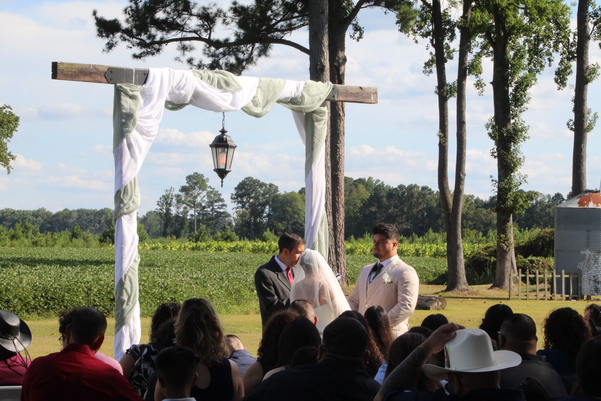 Wedding ceremony under a draped rustic wooden arch with a hanging lantern, officiant and groom standing with the veiled bride against a lush green farmland backdrop at Scarborough House.