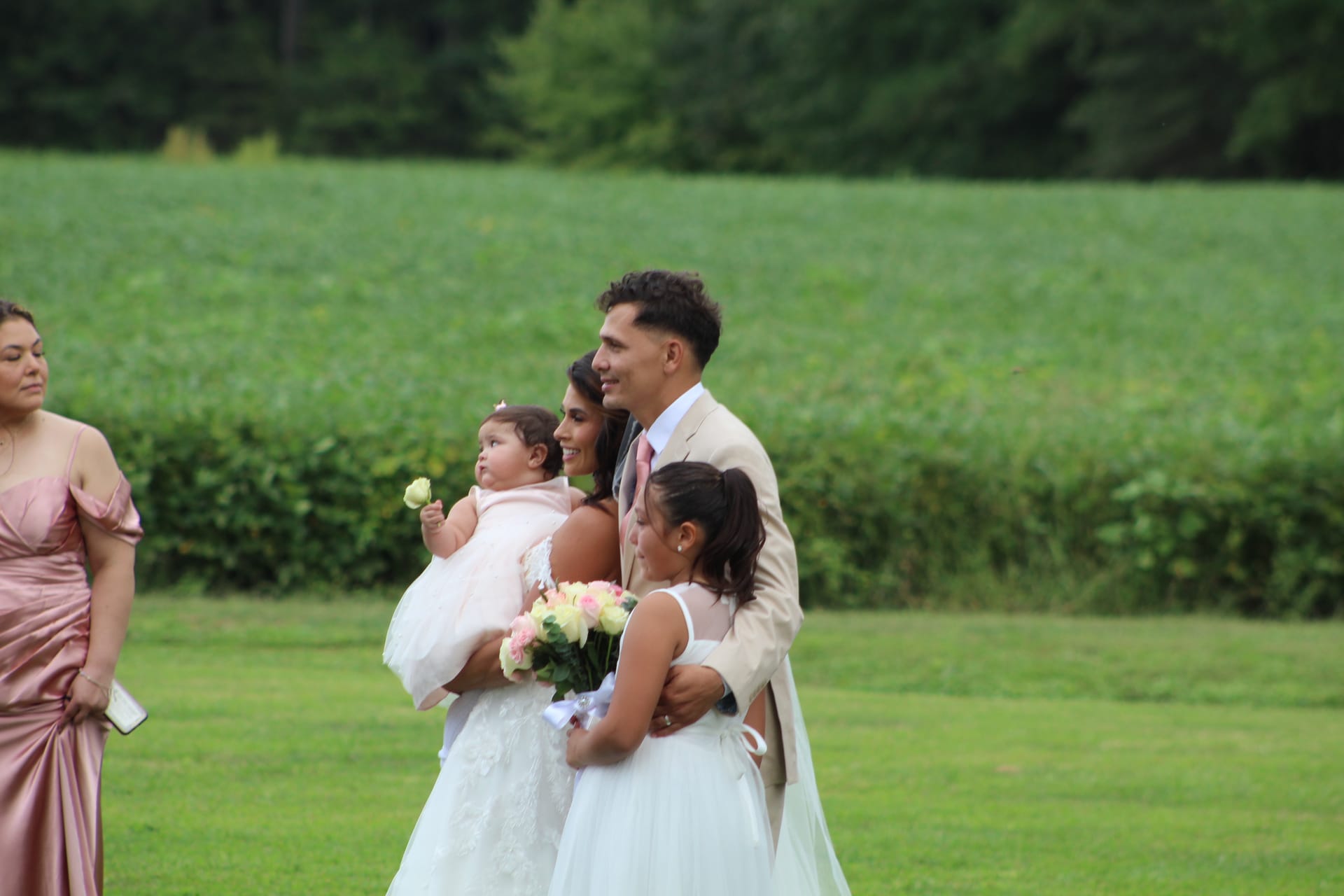 Smiling bride in a white gown holding a baby and a bouquet of pink roses stands beside the groom in a tan suit as a young flower girl leans in during an outdoor wedding ceremony at Scarborough House.