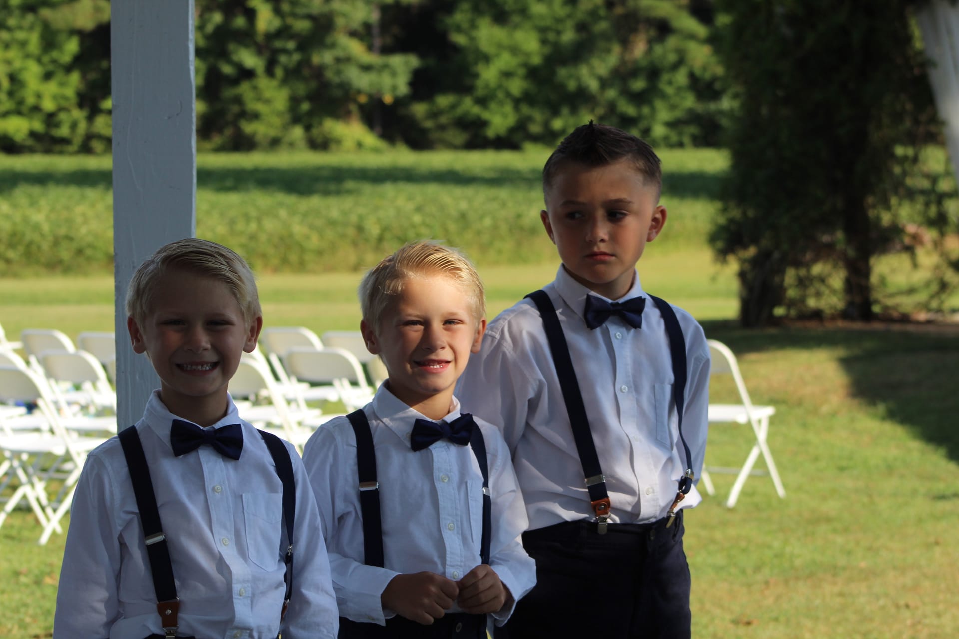 Three young ring bearers wearing white shirts, navy suspenders, and bow ties smile on the outdoor ceremony lawn at Scarborough House wedding venue.