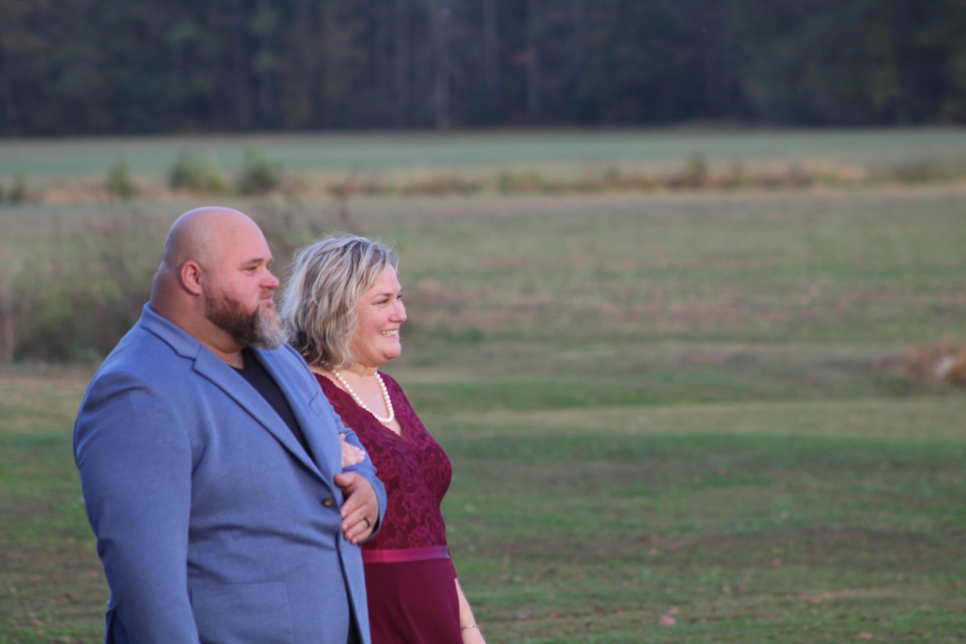 A smiling couple dressed in burgundy and blue walk arm-in-arm through open farmland fields at golden hour on the Scarborough House estate.