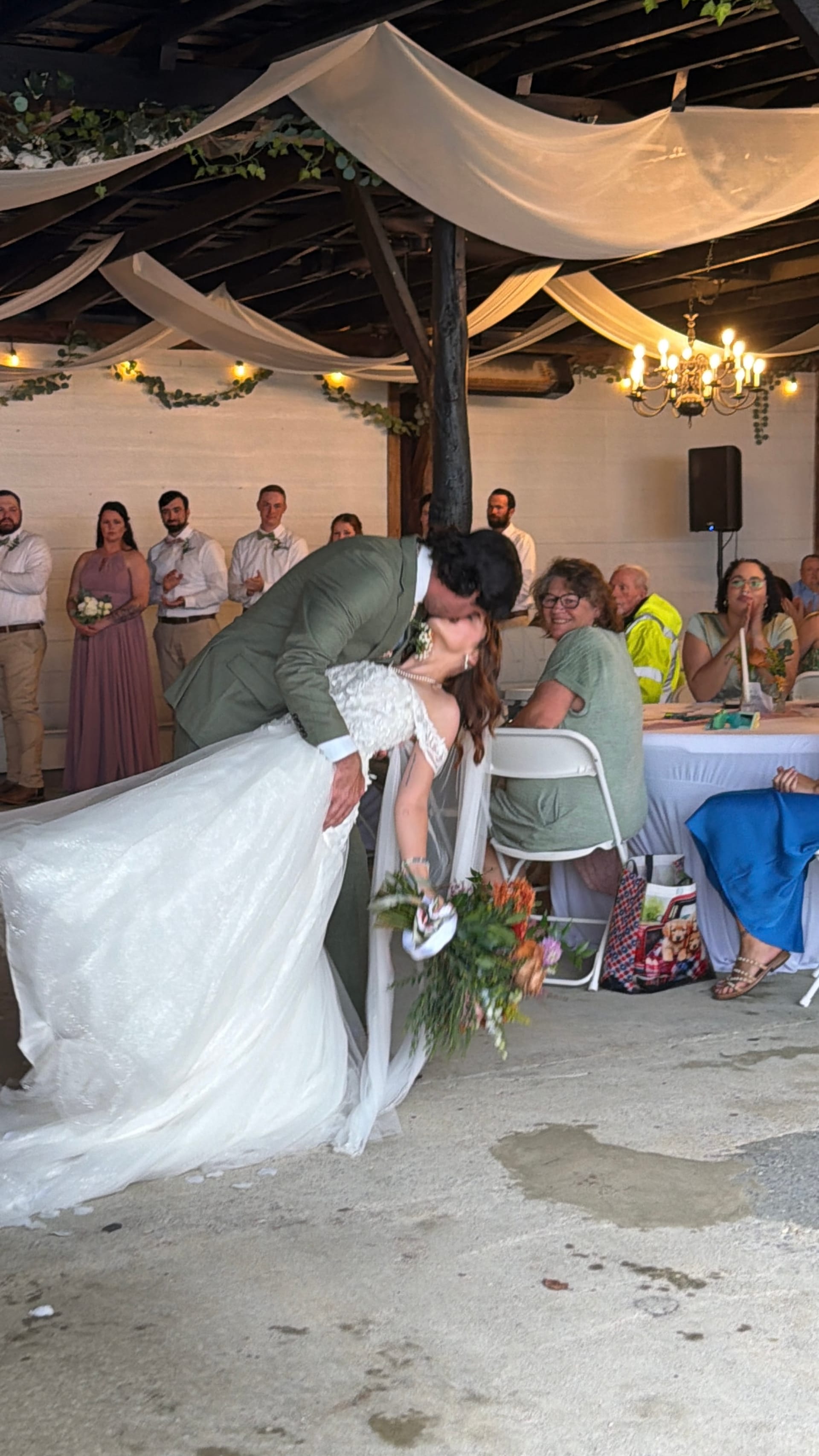 Groom in sage green suit dips and kisses his bride in a flowing white lace gown during their first dance inside a draped pavilion while guests and the wedding party look on.
