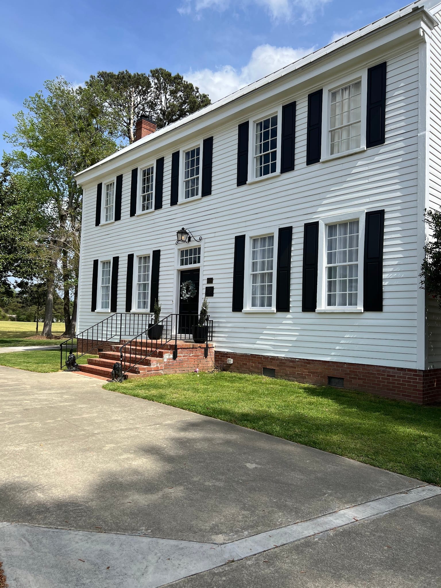Front facade of the historic 1821 Scarborough House manor featuring white clapboard siding, black shutters, brick entry steps, and manicured lawn under a blue sky.