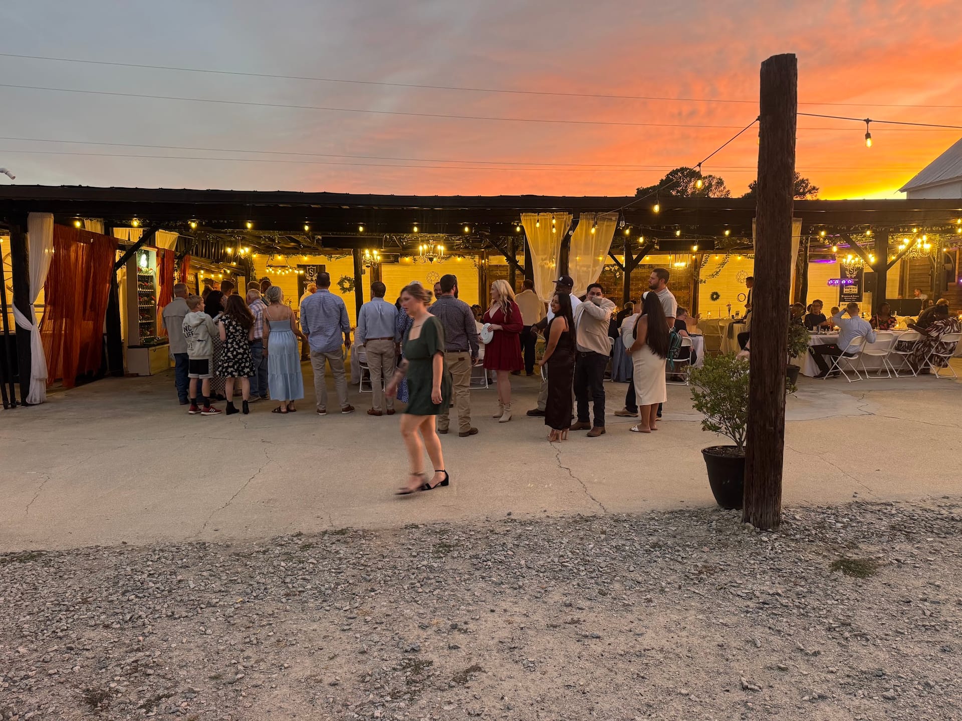 Wedding guests mingle under a string-lit outdoor pavilion at Scarborough House during a vibrant orange and pink sunset.