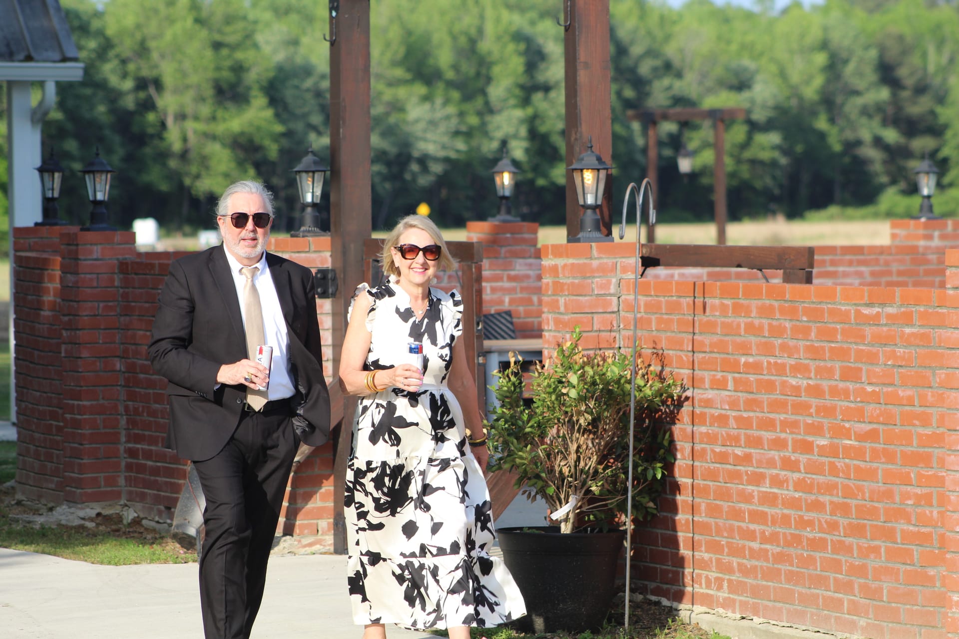 A well-dressed couple in formal attire walks smiling past the brick entrance wall with lantern posts at Scarborough House wedding venue on a sunny day.