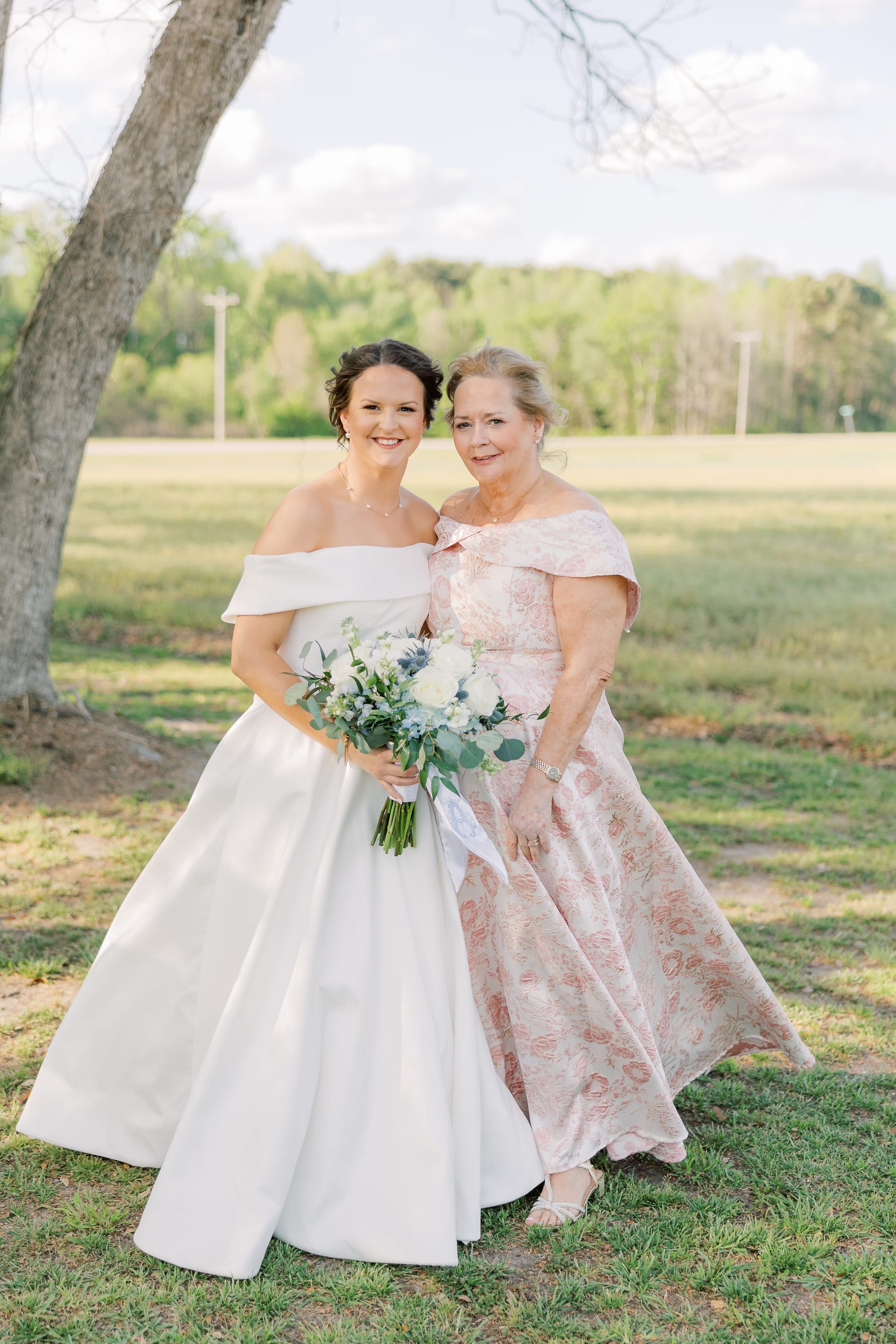 Bride in an off-shoulder white ball gown holding a greenery and white floral bouquet poses with an older woman in a pink floral off-shoulder gown beneath oak trees on a sunny day at Scarborough House.