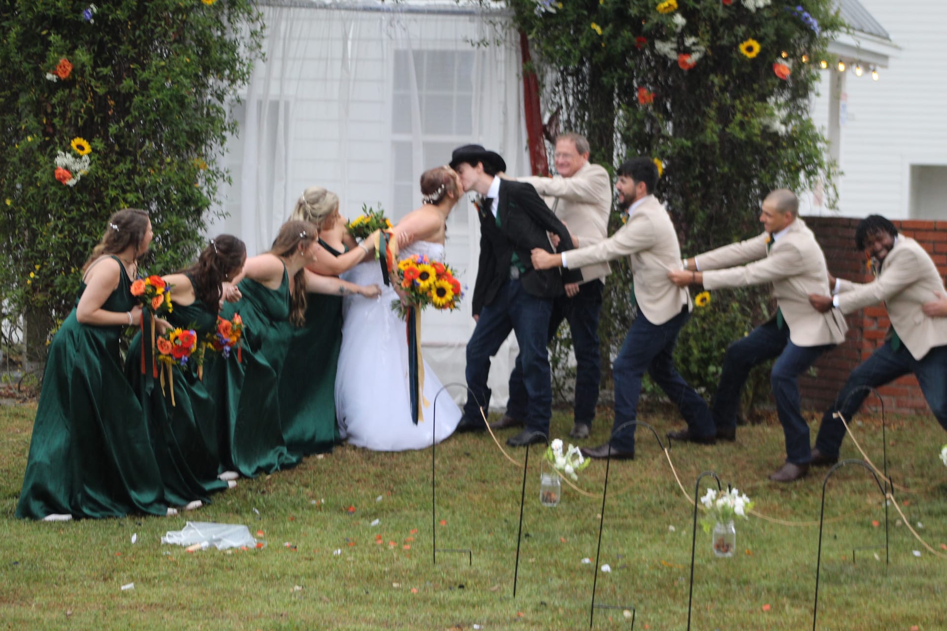 Bridesmaids in emerald green gowns and groomsmen in tan blazers playfully pull the bride and groom apart during their kiss on the ceremony lawn at Scarborough House historic estate.