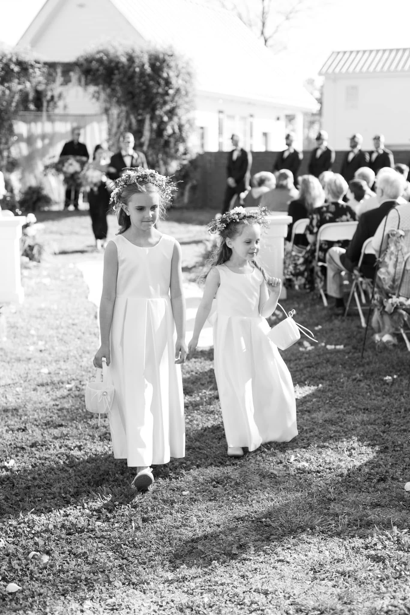 Two young flower girls in white dresses and floral crowns walk hand-in-hand down the outdoor ceremony aisle at Scarborough House while seated guests look on.