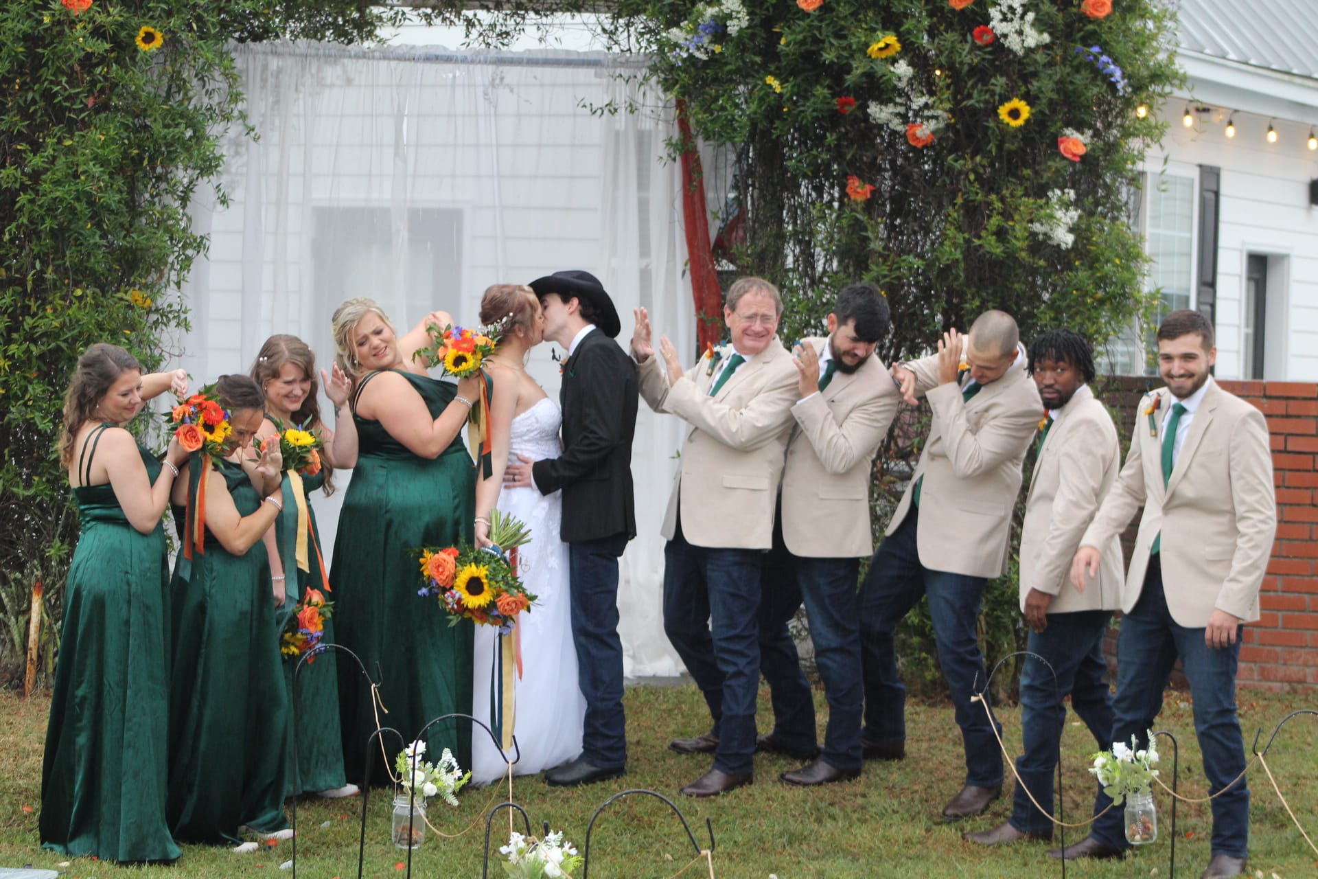 Bride and groom share their first kiss at a sunflower-adorned arch on the ceremony lawn of Scarborough House as bridesmaids in emerald gowns and groomsmen in tan suits celebrate around them.