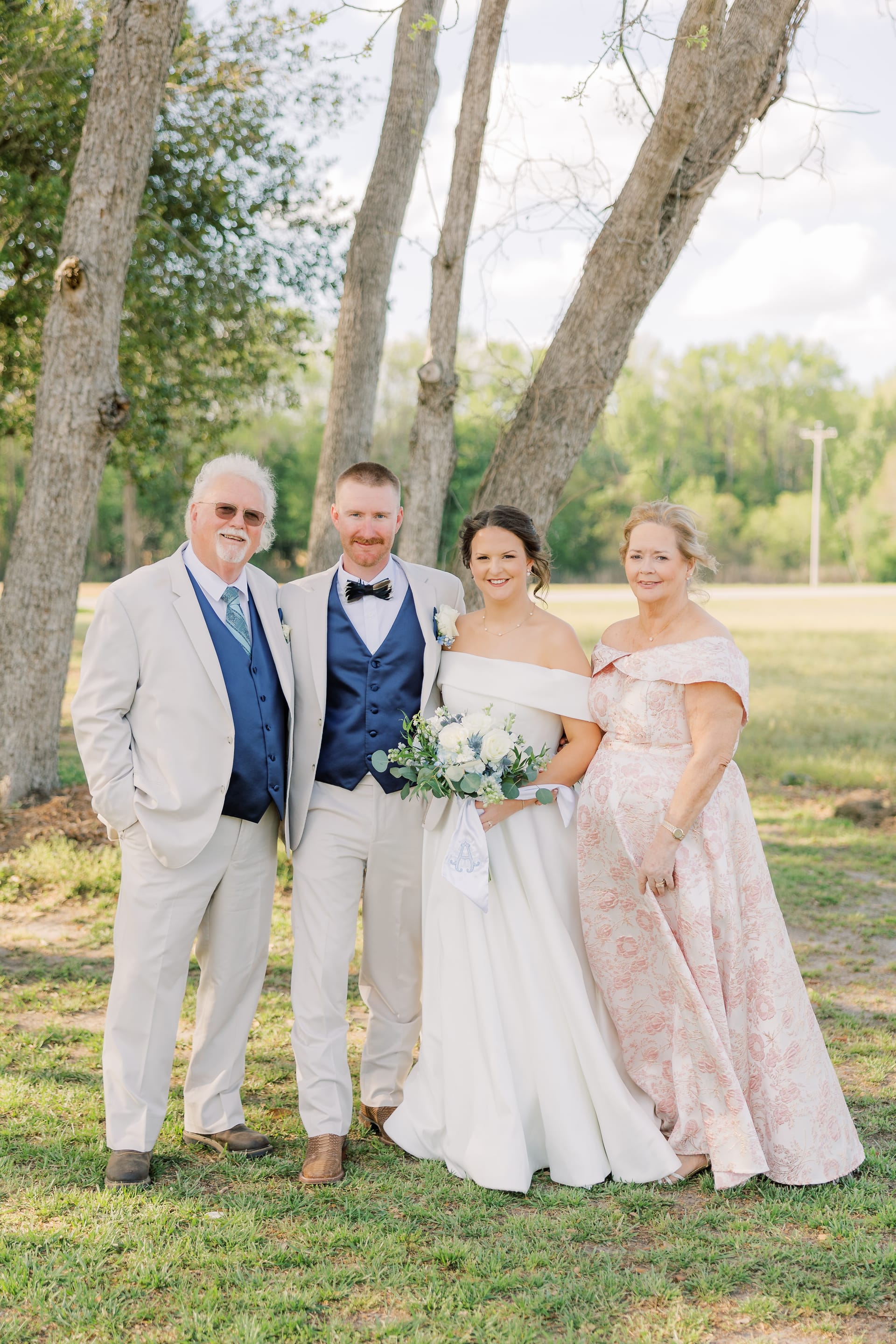 Bride and groom pose with parents under large oak trees on the grounds of Scarborough House, the bride holding a white and greenery bouquet while wearing an off-shoulder gown and the groom a navy vest with bow tie.