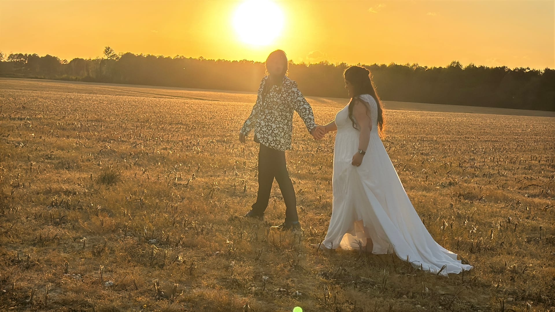 Bride in a flowing white gown and groom in a floral shirt walk hand in hand through golden farmland fields at Scarborough House as the sun sets behind them.