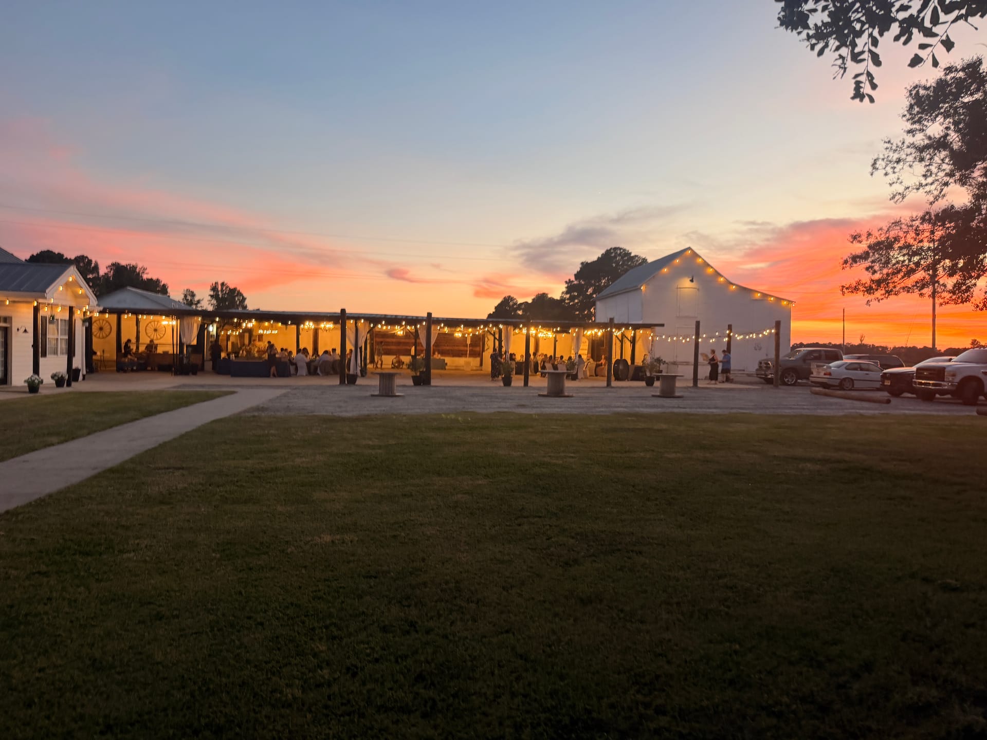 Warm string lights illuminate an outdoor covered reception pavilion at Scarborough House against a vivid pink and orange sunset sky with guests gathered inside.