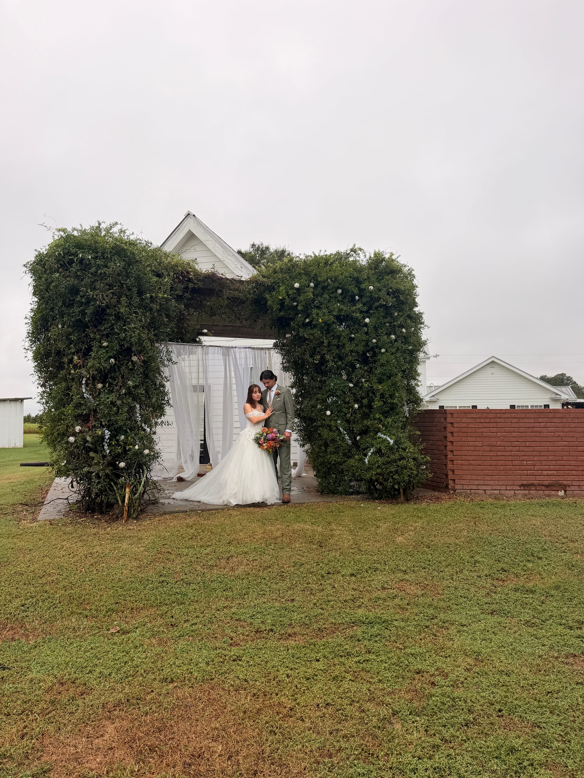 Bride in a white ball gown and groom in a grey suit pose together under a large ivy-covered arch with white draping at Scarborough House estate wedding venue.