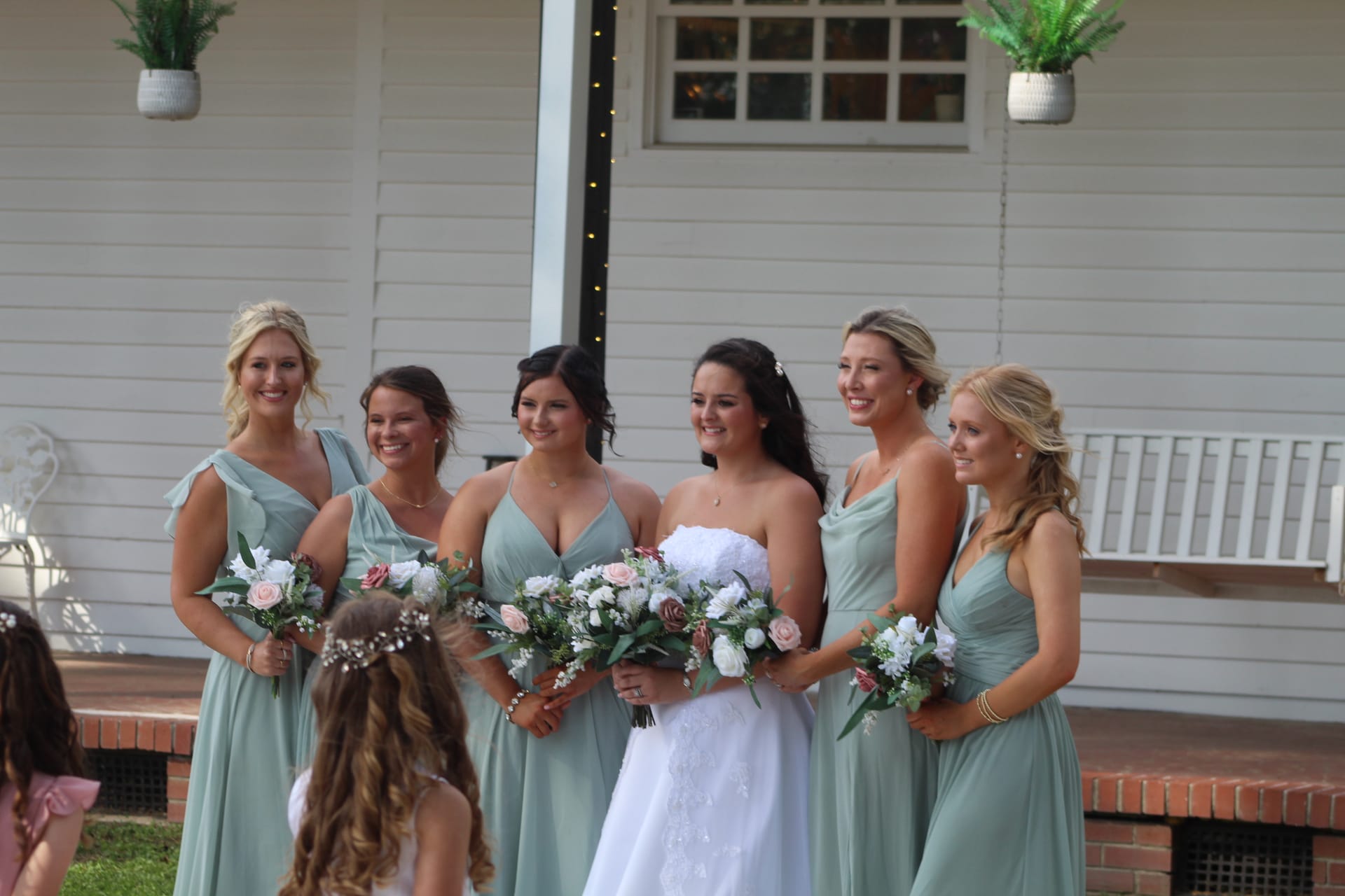 Bride in strapless white gown stands smiling with five bridesmaids in sage green dresses holding floral bouquets in front of the white clapboard facade of Scarborough House historic estate.