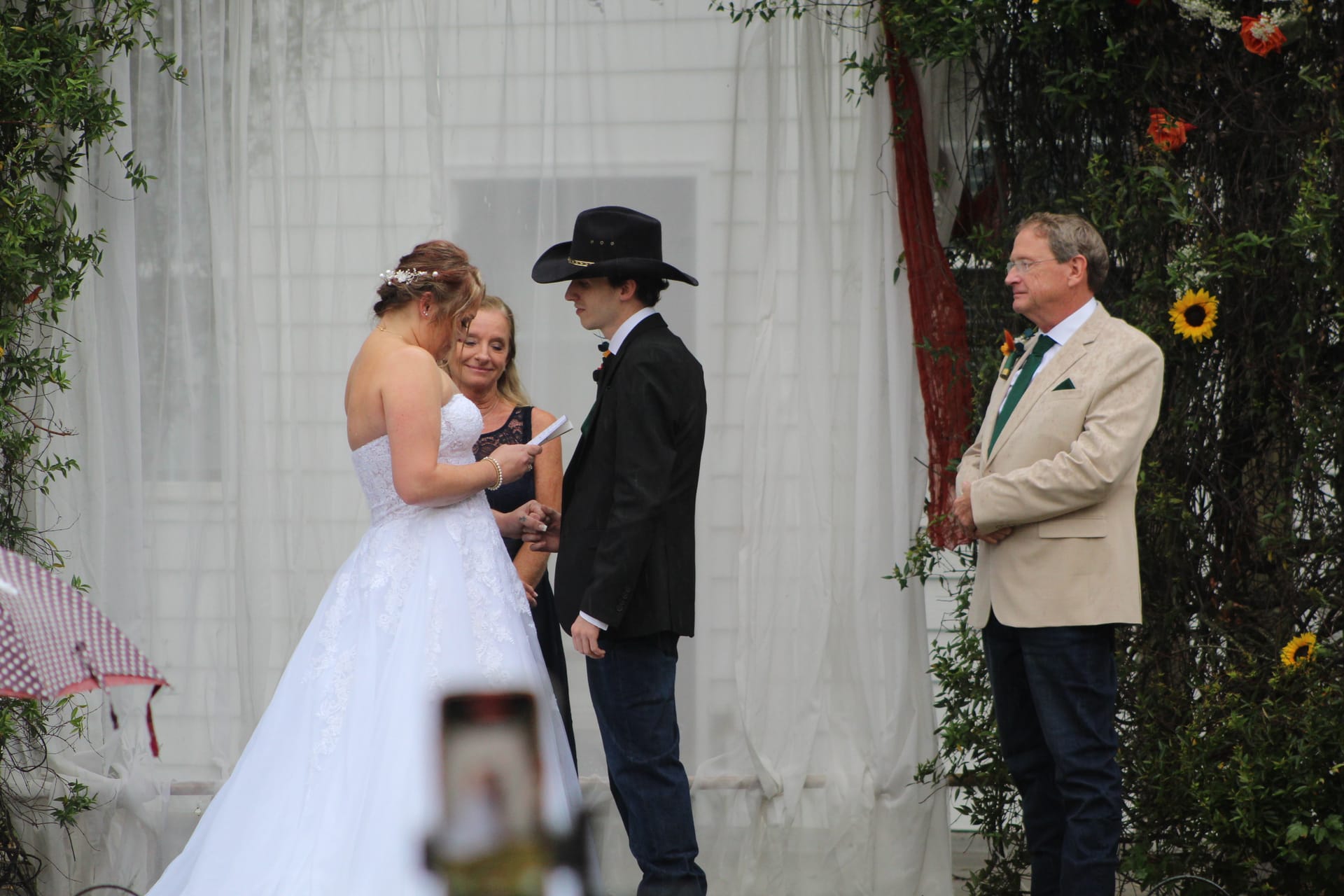 Bride in a white ball gown reads vows to groom in a black cowboy hat and blazer beneath a lush floral arch at Scarborough House outdoor ceremony.