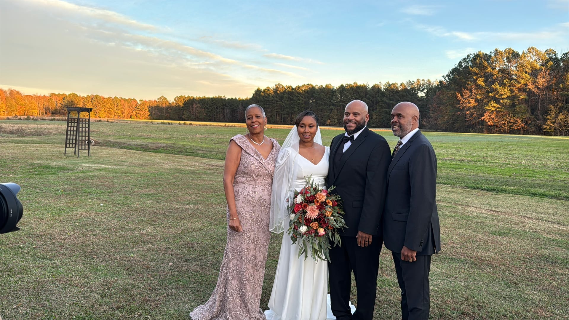 Bride in white gown holding a colorful cascading bouquet poses with three family members in formal attire on the open ceremony lawn at golden hour with autumn trees in the background.