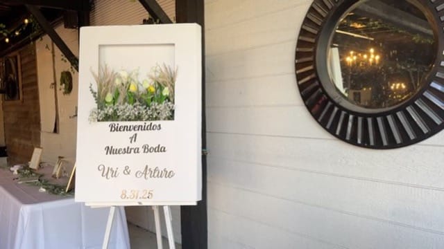 White framed wedding welcome sign reading Bienvenidos A Nuestra Boda for Uri and Arturo on 8.31.25 adorned with yellow tulips and baby's breath on a wooden easel inside Scarborough House.