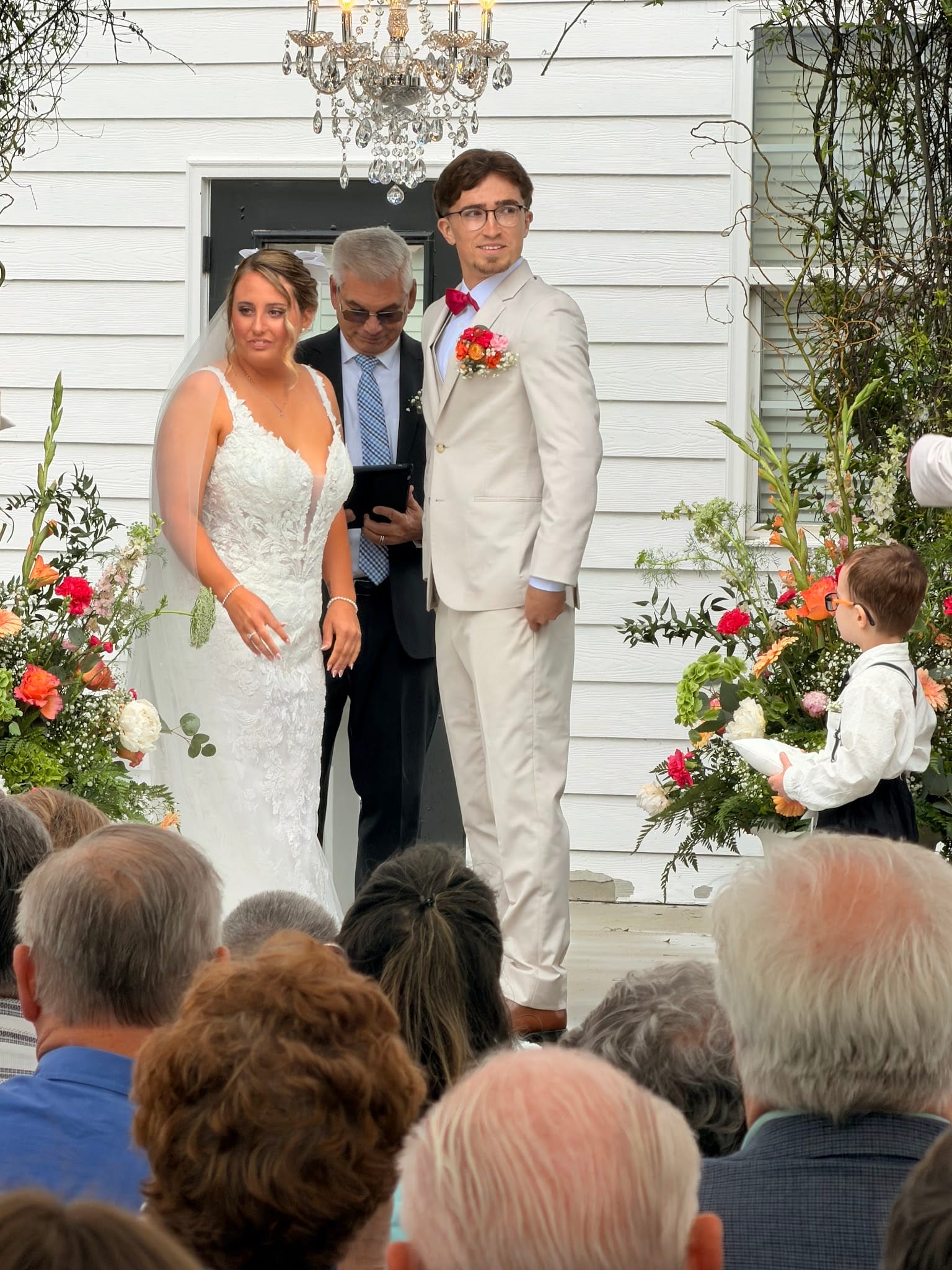 Bride in lace gown and groom in cream suit stand with officiant during outdoor wedding ceremony at a white historic manor adorned with vibrant floral arrangements and a crystal chandelier.