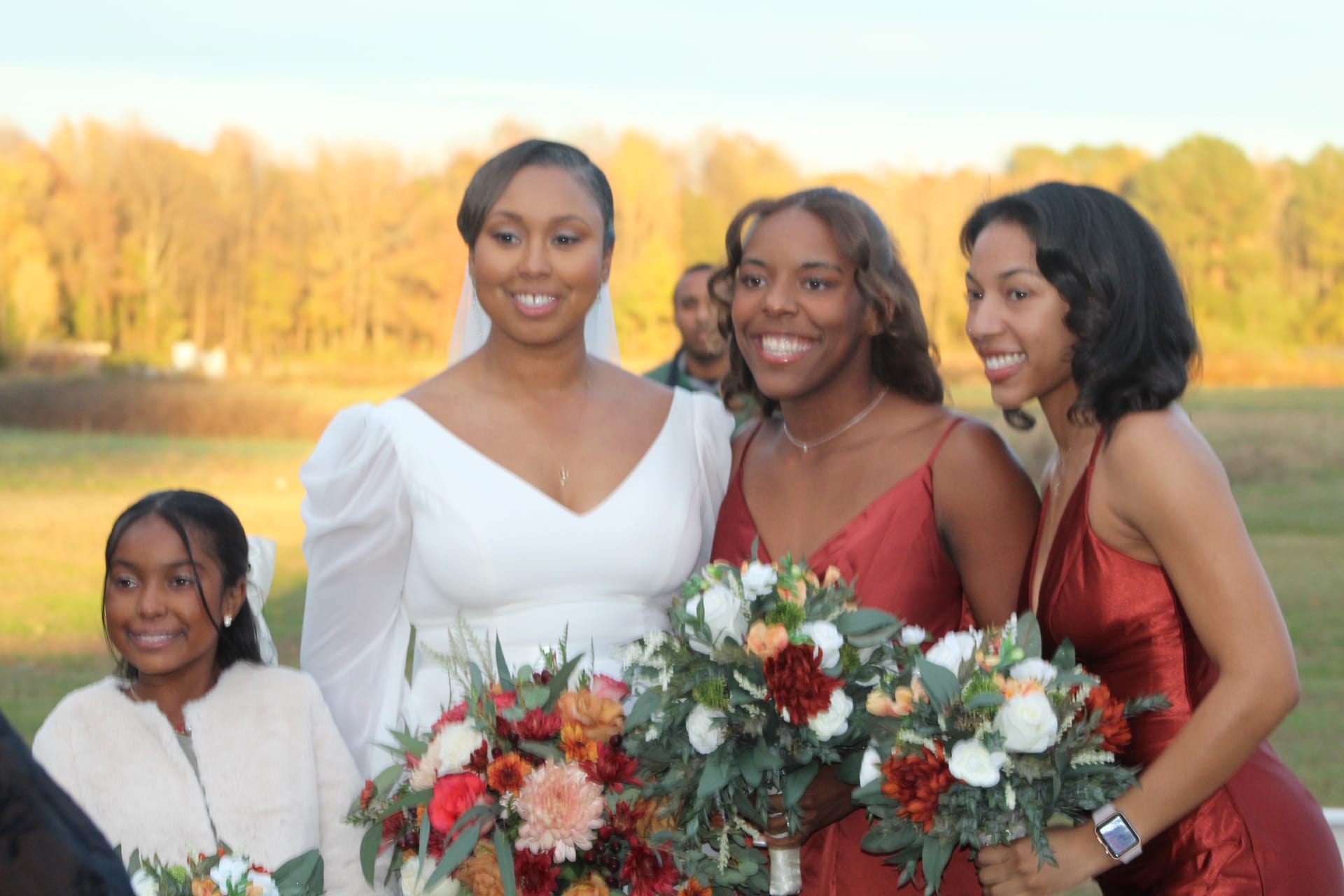 Bride in a white long-sleeve gown stands with two bridesmaids in rust-red satin dresses holding lush autumn bouquets of dahlias and roses, with golden fall foliage in the background at Scarborough House.