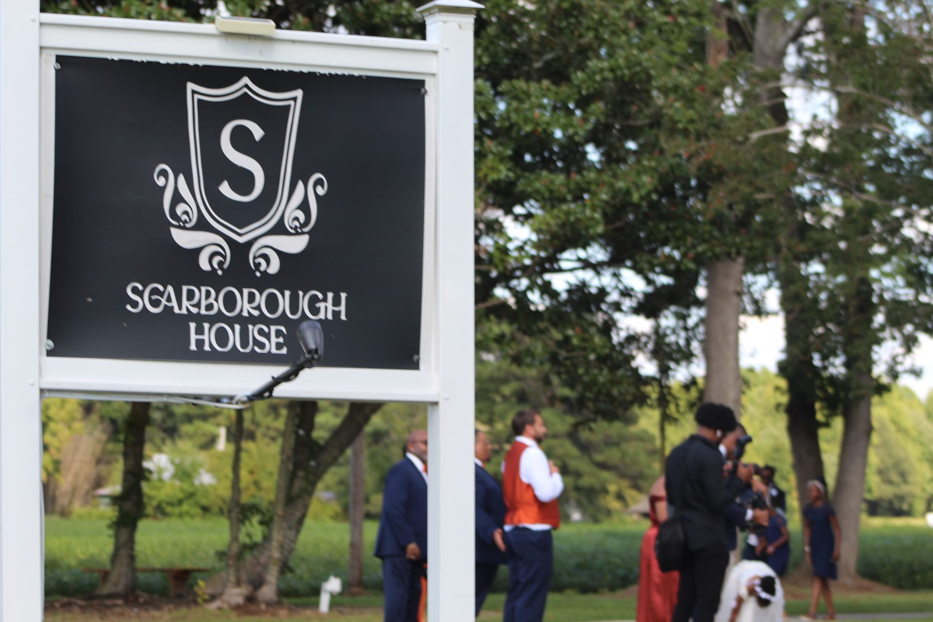 Scarborough House estate wedding venue sign in the foreground with formally dressed guests mingling among mature oak trees and manicured grounds in the background.