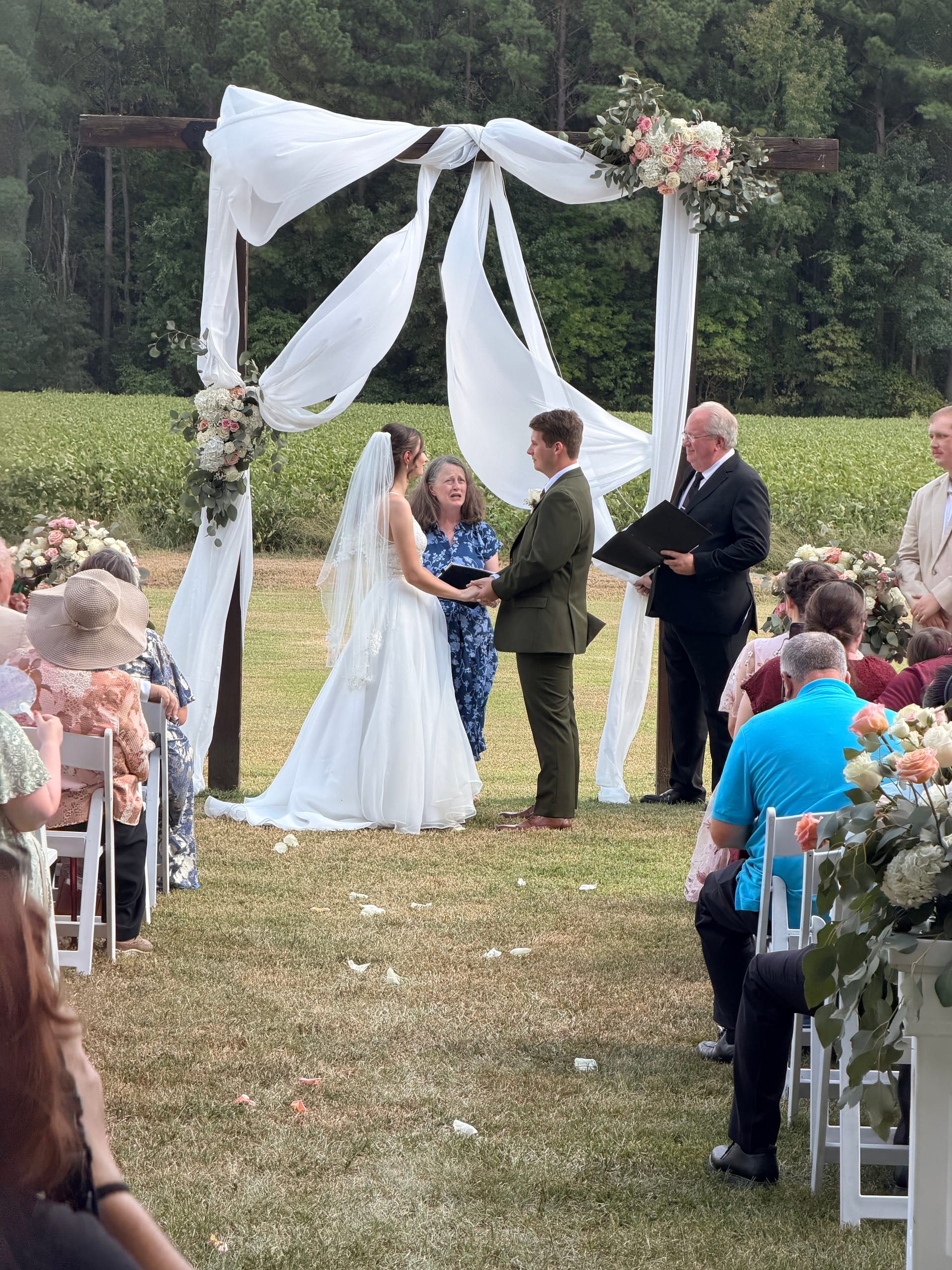 Bride and groom exchange vows beneath a white-draped wooden arch adorned with blush floral arrangements on the ceremony lawn at Scarborough House, surrounded by seated guests and lush farmland fields.