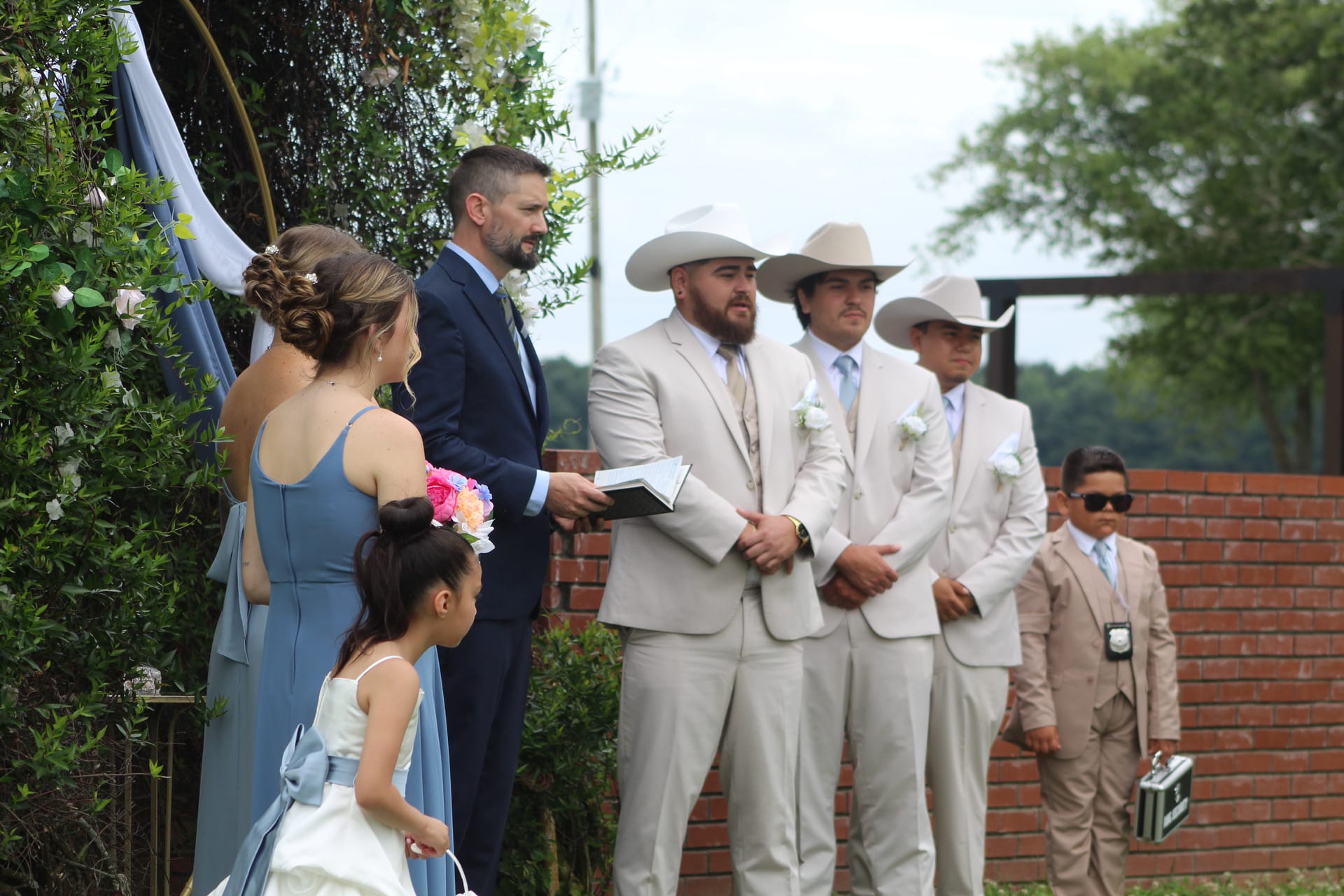 Groomsmen in tan suits and cowboy hats stand at attention during an outdoor wedding ceremony at Scarborough House, with a flower girl and bridesmaid visible near a floral arch.