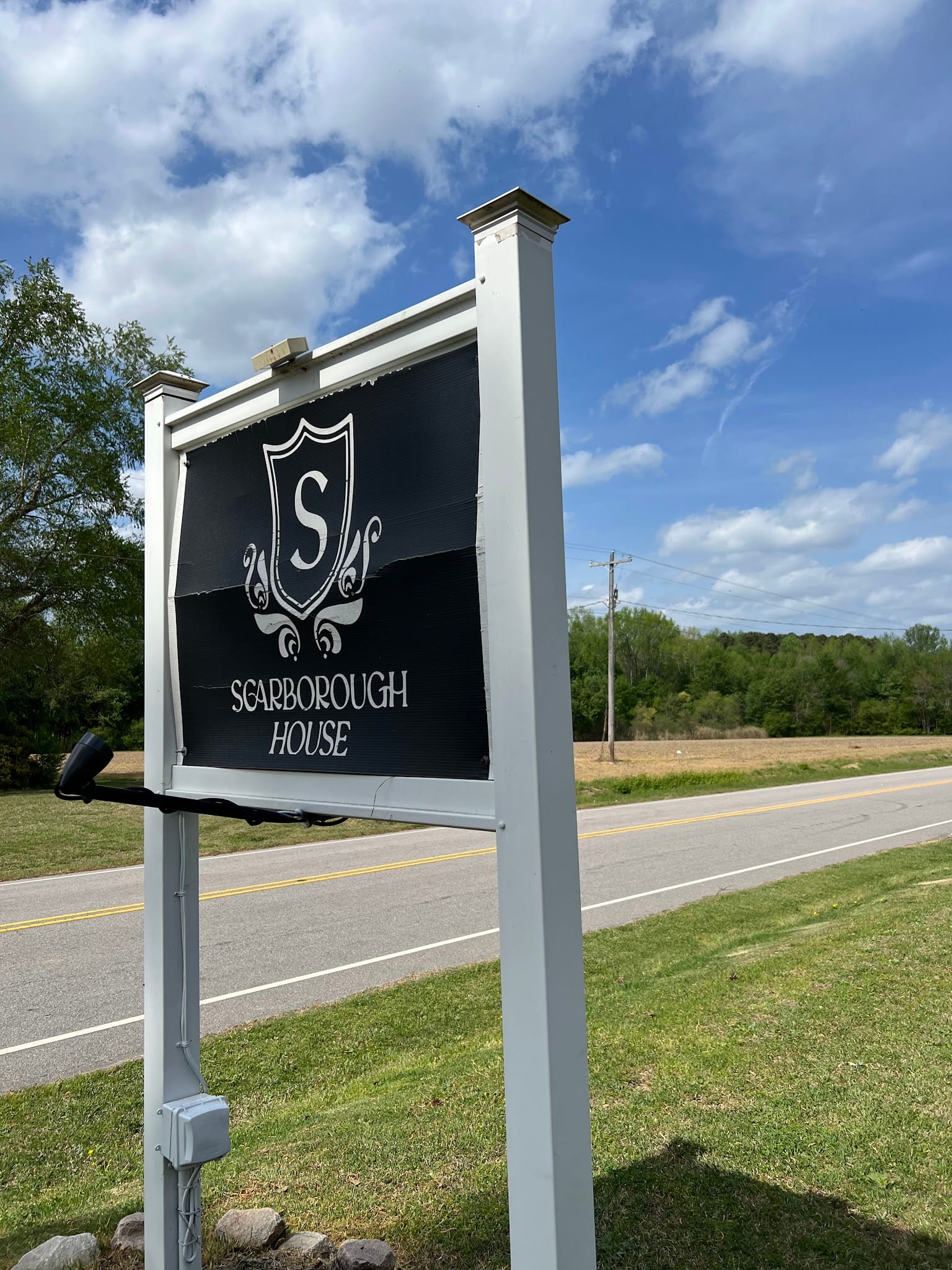 Black and white Scarborough House estate sign mounted on white posts along a tree-lined country road under a partly cloudy blue sky.