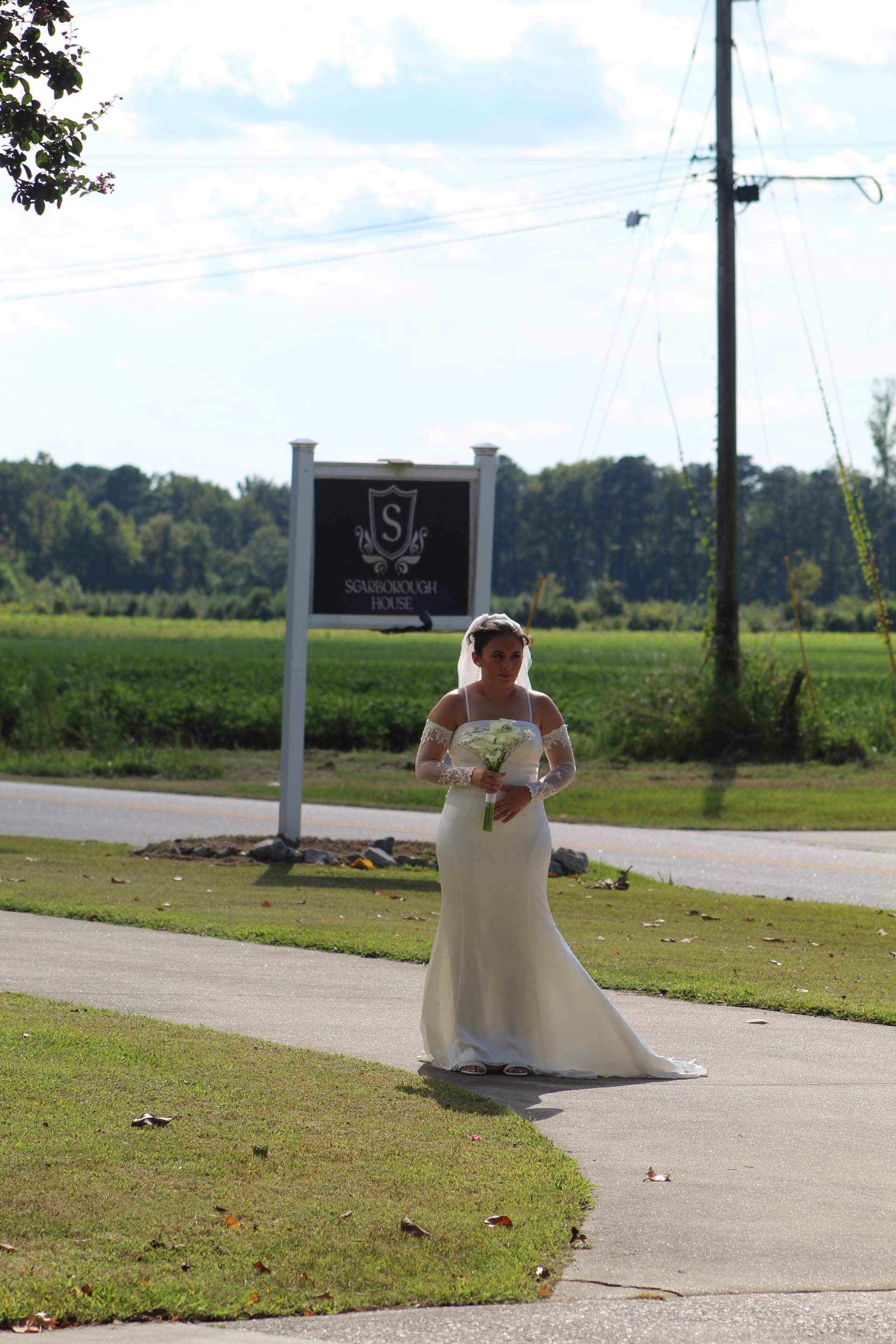 Bride in a white off-shoulder gown holding a white floral bouquet stands near the Scarborough House entrance sign with lush green fields and trees in the background.