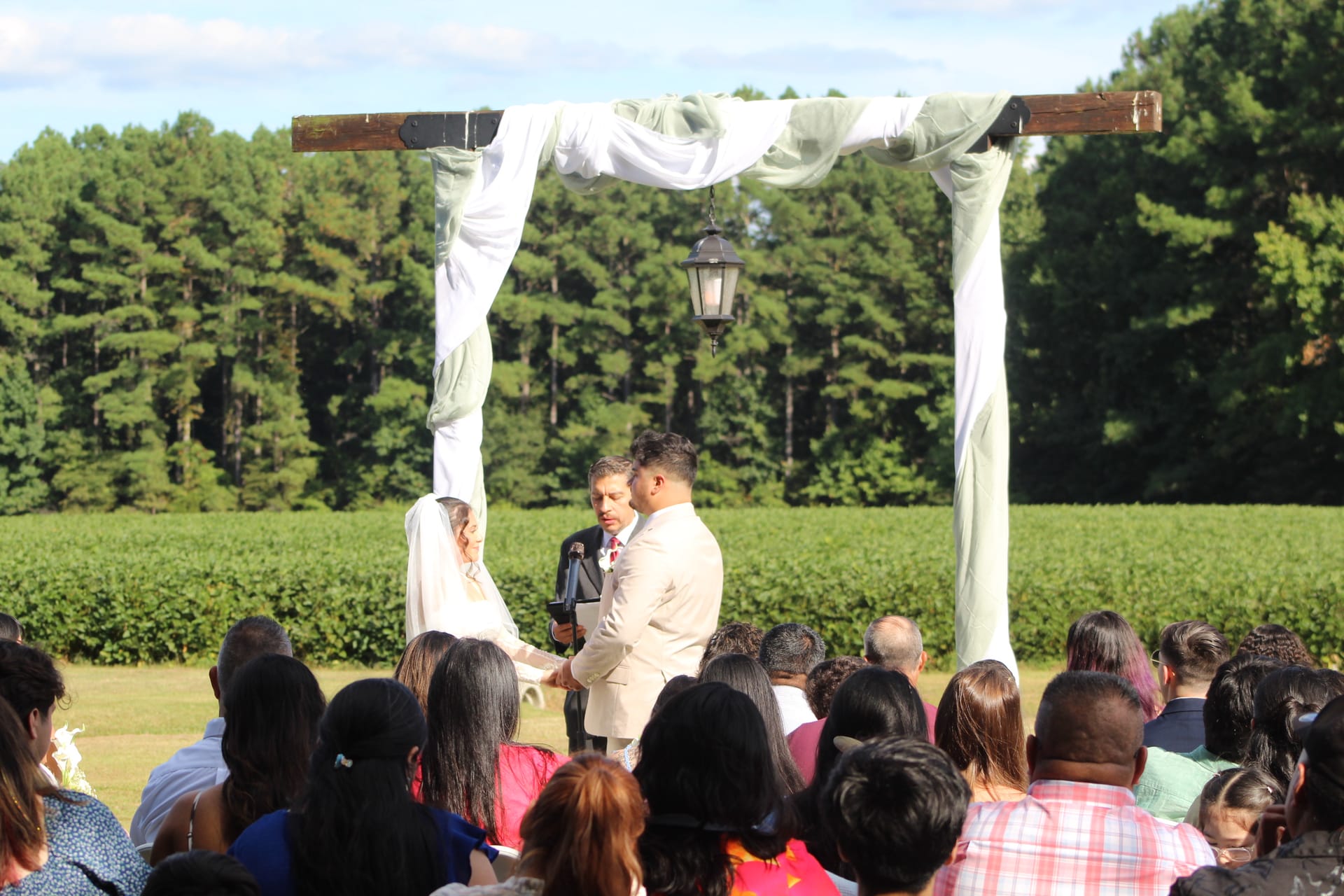 A bride and groom exchange vows beneath a draped wooden arch lantern on the ceremony lawn at Scarborough House, surrounded by seated guests with lush green fields and trees in the background.