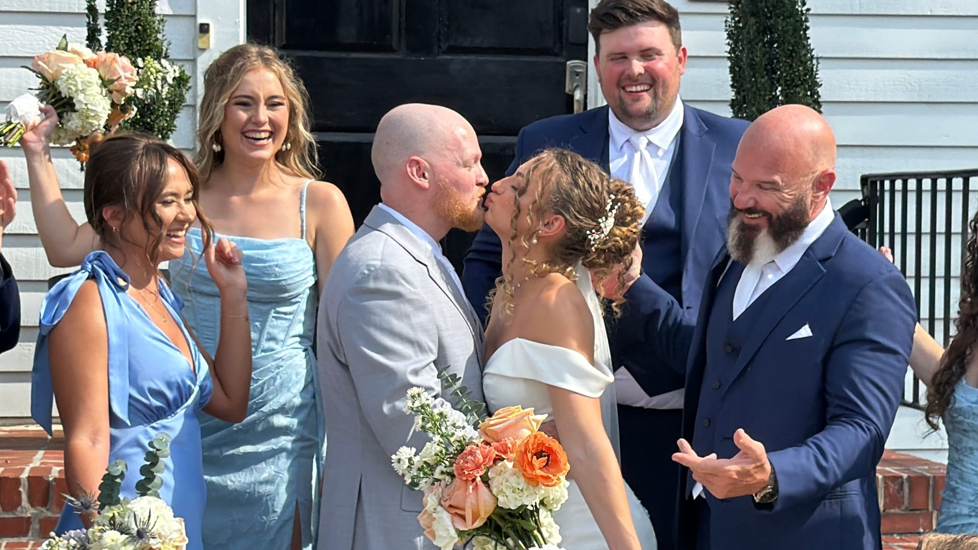 Bride and groom share their first kiss on the front steps of Scarborough House as smiling bridesmaids in blue dresses and groomsmen in navy suits celebrate around them.