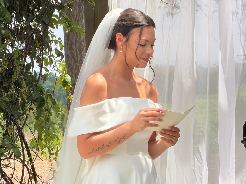 Bride in an off-shoulder white gown and cathedral veil reads her handwritten vows during an outdoor ceremony at Scarborough House, surrounded by lush greenery and soft natural light.