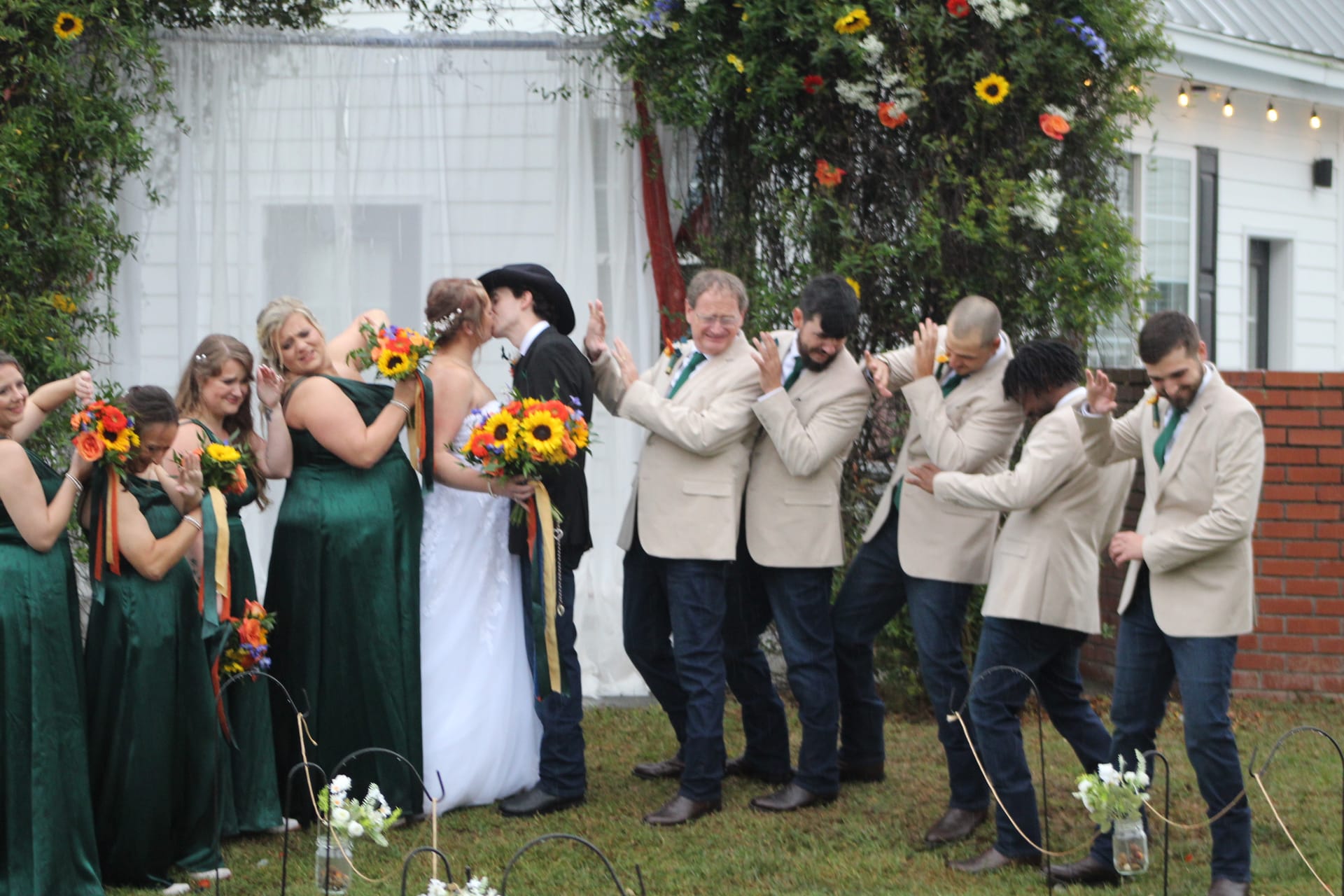 Bride and groom share their first kiss surrounded by cheering bridesmaids in emerald green dresses and groomsmen in cream blazers on the ceremony lawn at Scarborough House.