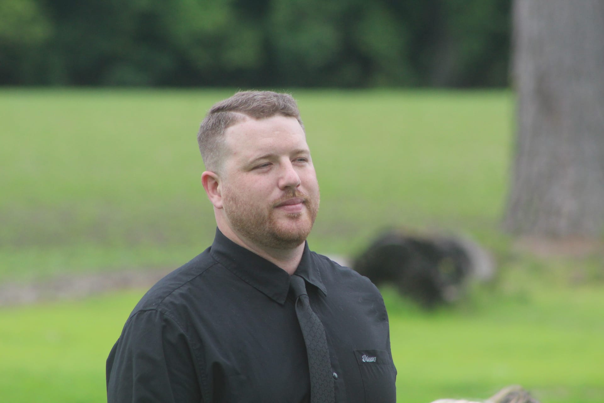 A man in a black dress shirt and tie smiles candidly on the ceremony lawn at Scarborough House estate wedding venue, with lush green fields and trees in the background.