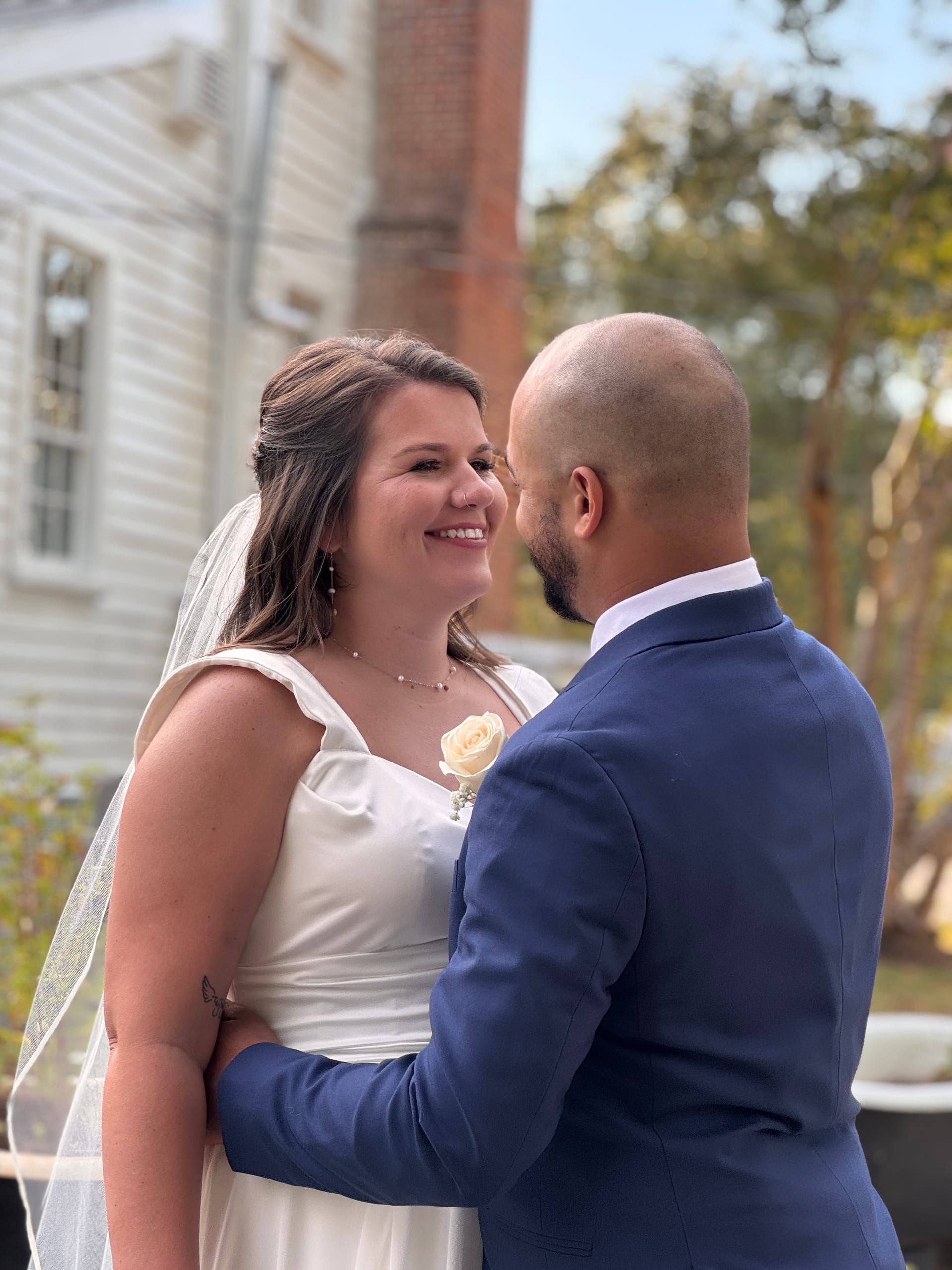 Smiling bride in a white gown and veil gazes lovingly at her groom in a navy suit during their outdoor wedding ceremony at the historic 1821 Scarborough House estate.