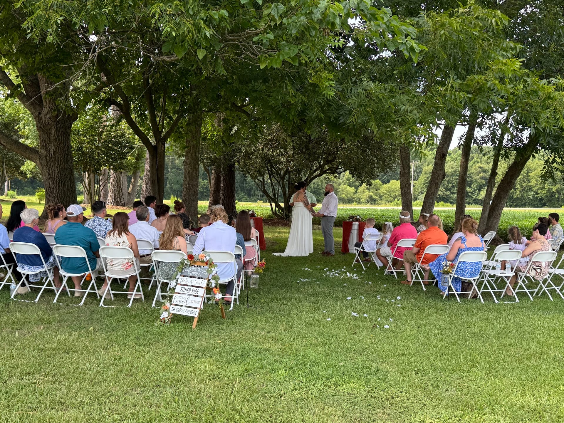 Guests seated in white folding chairs on a shaded lawn witness an outdoor wedding ceremony beneath tall canopy trees at Scarborough House historic estate.
