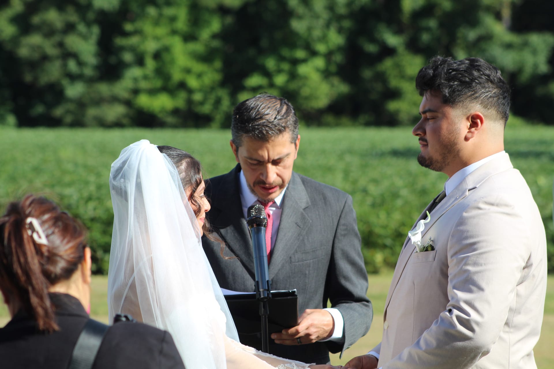Bride and groom exchange vows during an outdoor wedding ceremony on the sunlit ceremony lawn, with an officiant reading from a book and green farmland fields in the background.