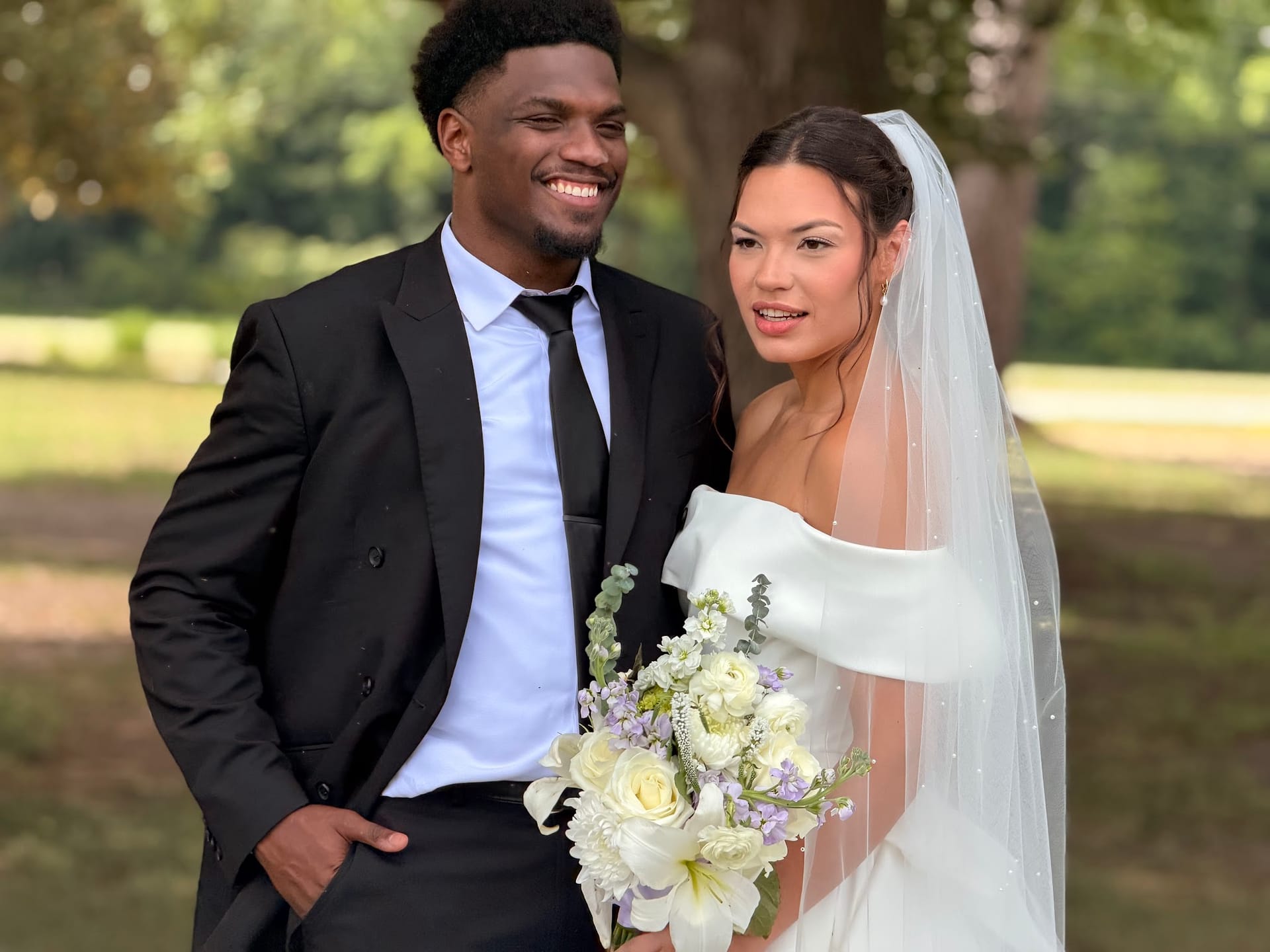 Smiling groom in a black suit and bride in an off-shoulder white gown holding a white and lavender bouquet pose together beneath oak trees at Scarborough House estate.