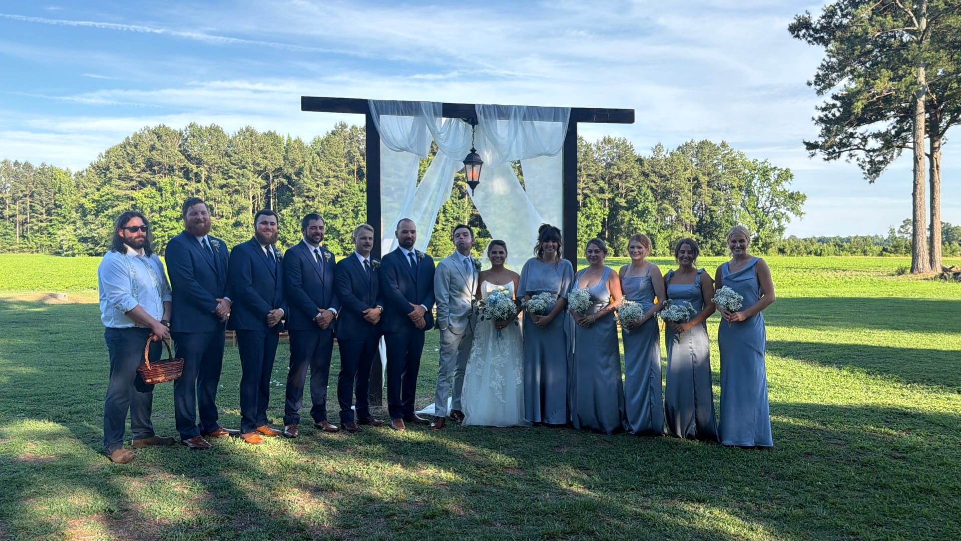 Bride and groom stand at the center of their wedding party in front of a draped wooden ceremony arch on the sunlit ceremony lawn at Scarborough House, surrounded by groomsmen in navy suits and bridesmaids in dusty blue gowns holding white floral bouquets.