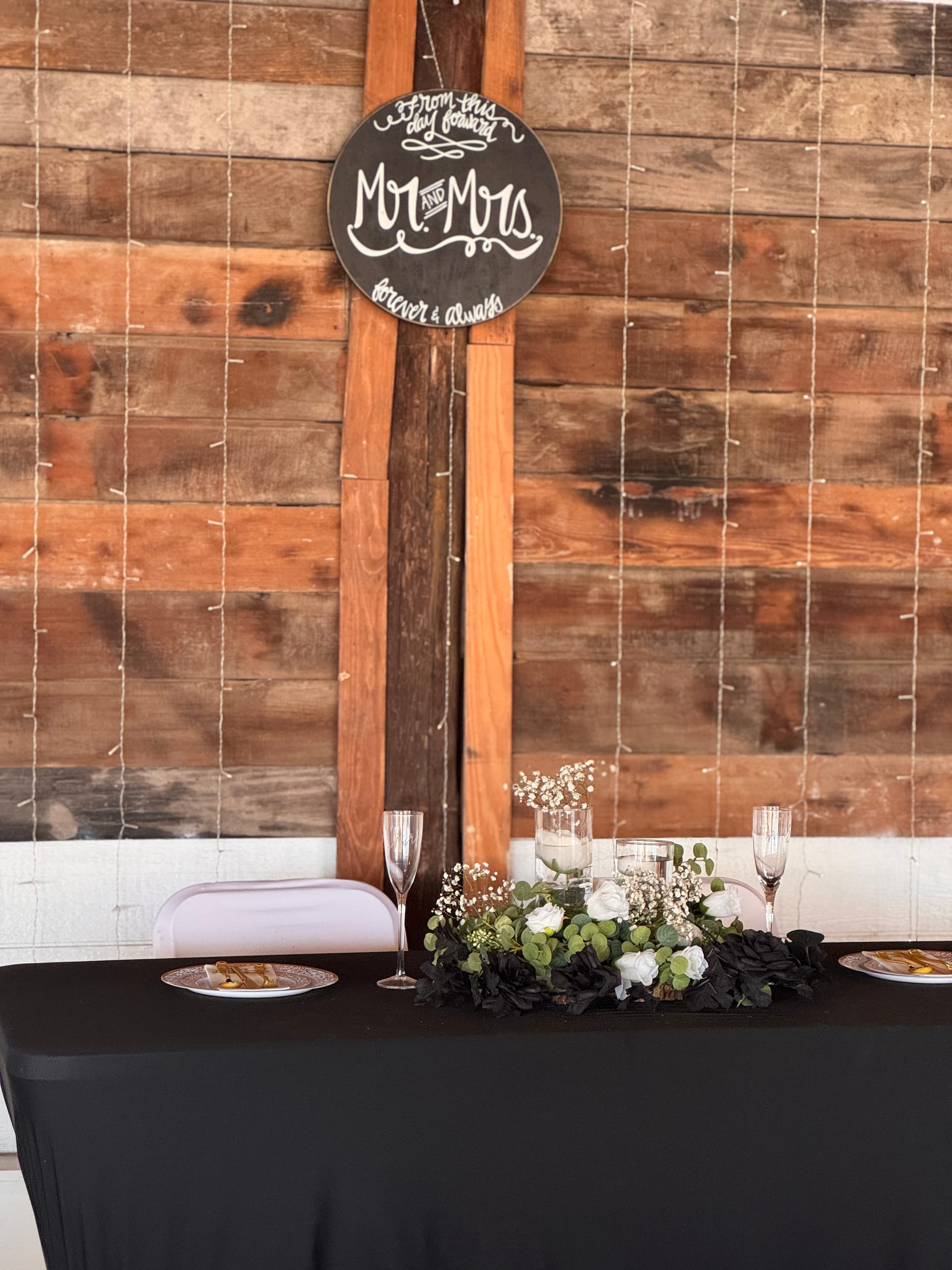 Rustic sweetheart table draped in black linen with white and dark floral centerpiece, champagne flutes, and a chalkboard Mr and Mrs sign hanging on a reclaimed wood wall at Scarborough House wedding venue.
