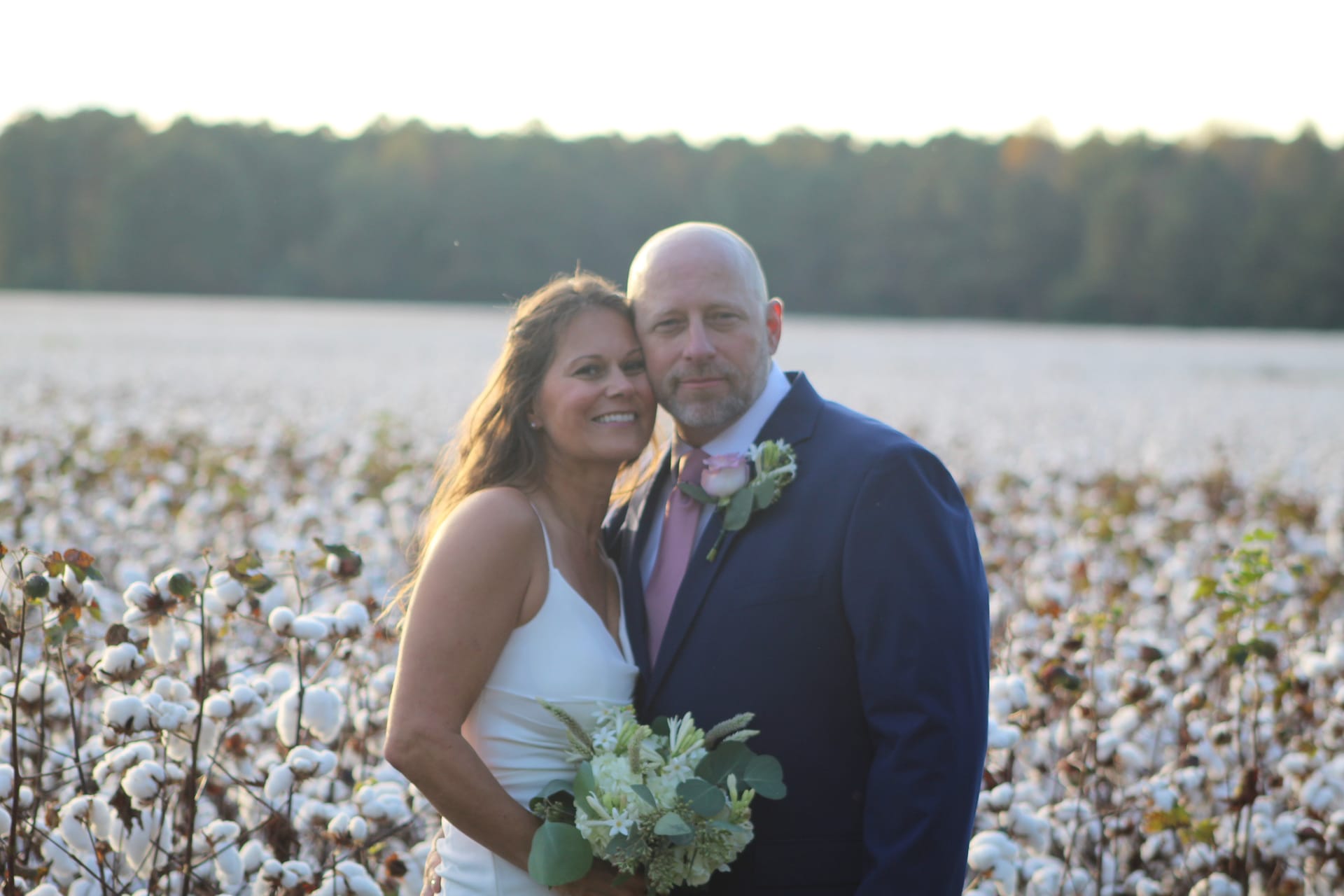 Bride in white gown holding a white floral bouquet and groom in a navy suit with pink tie and boutonniere pose together smiling in a cotton field at golden hour with a treeline in the background.