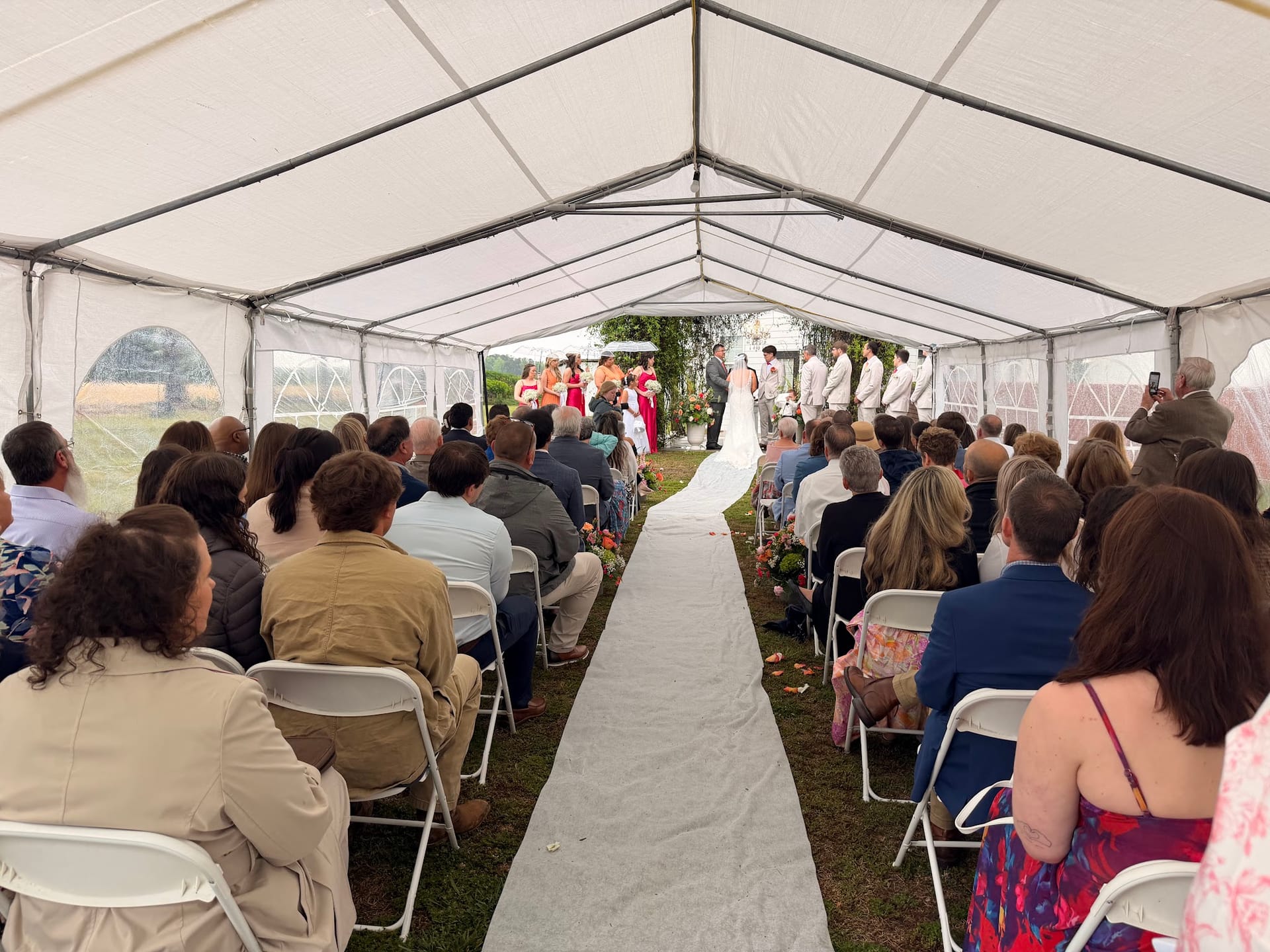 Guests seated in white folding chairs beneath a large white event tent watch a wedding ceremony at the far end of a white aisle runner at Scarborough House.