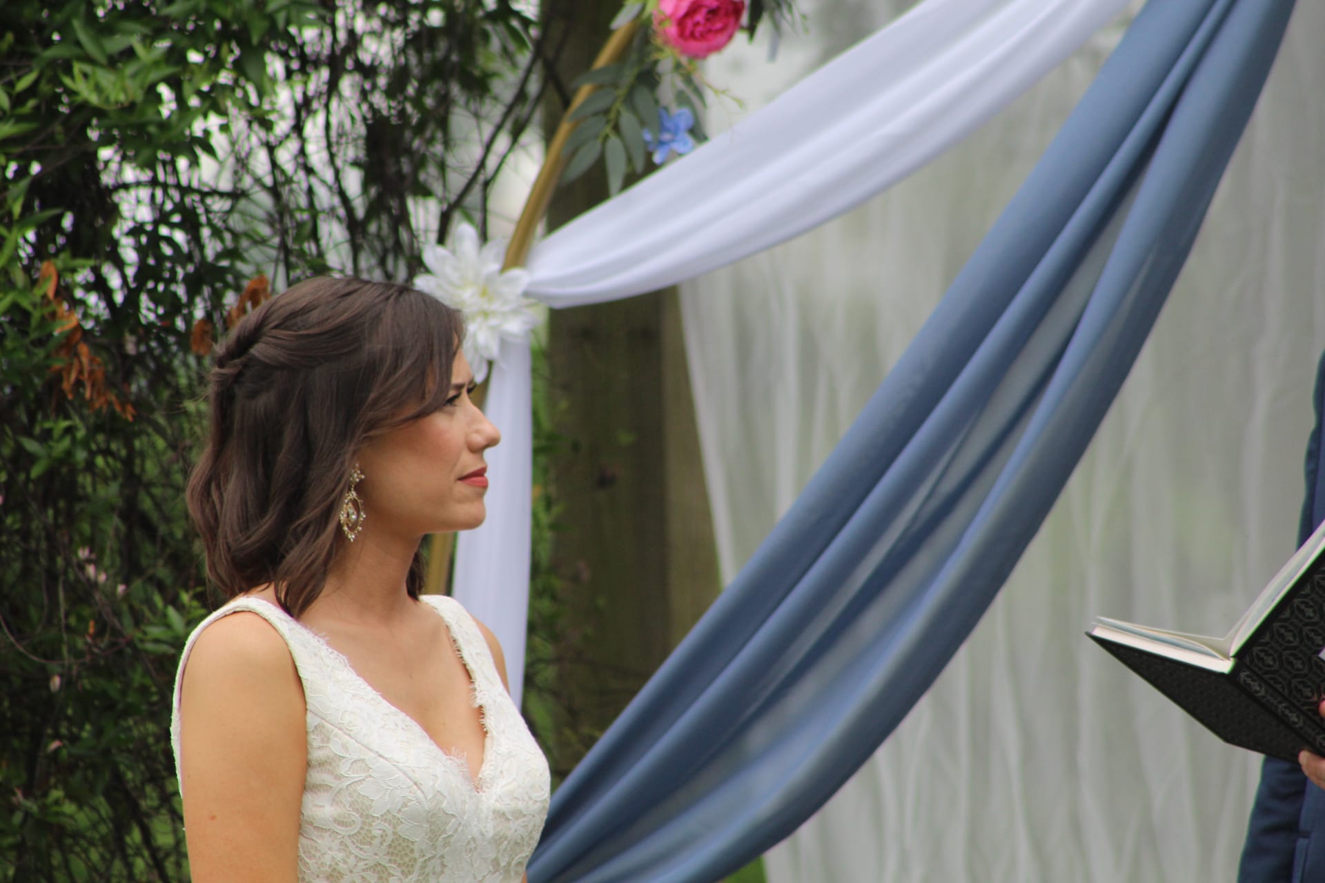 Bride in a lace sleeveless gown gazes forward during her outdoor wedding ceremony beneath a draped arch adorned with blue fabric and pink florals at a historic estate venue.