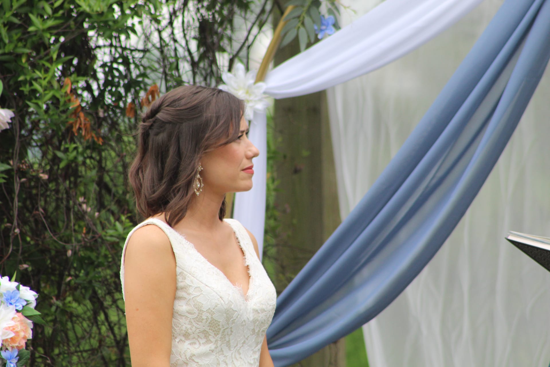Bride in a sleeveless lace gown gazes forward during an outdoor garden ceremony beneath a draped arch adorned with white and blue fabric at Scarborough House.